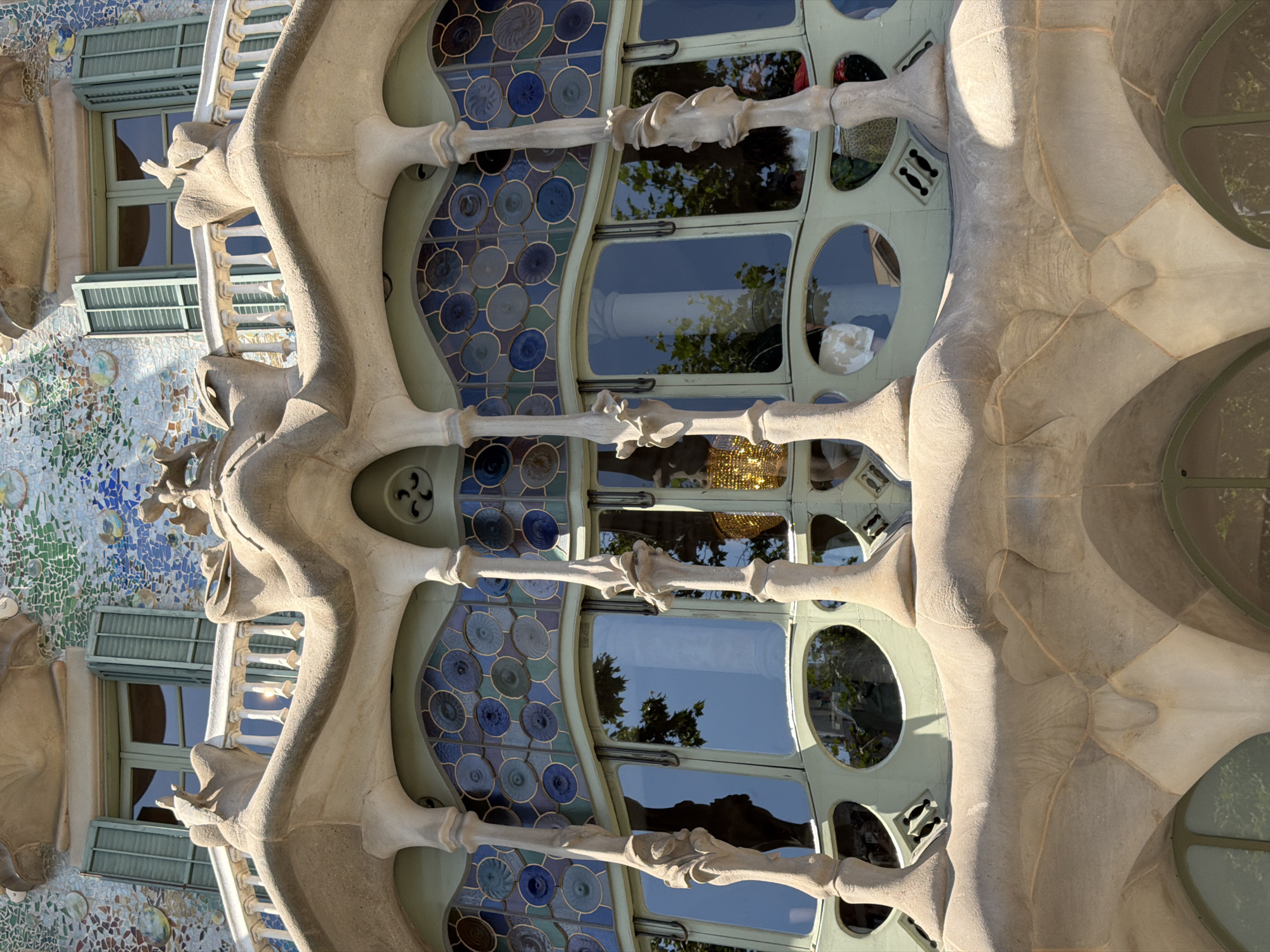 The image depicts a close-up, low-angle view of the facade of Casa Batlló, a building located in Barcelona, Spain, designed by Antoni Gaudí. The architecture is characterized by its organic, flowing forms and vibrant, mosaic-like ornamentation.
In the foreground, a balcony with an intricately sculpted, bone-like balustrade juts out from the building. The balustrade is made of a light-colored, textured material, possibly stone or concrete, and features a series of rounded elements and decorative protrusions. Supporting this balcony are several slender, column-like structures that also exhibit a fluid, somewhat skeletal appearance.
Behind the balcony, a large window section is visible, characterized by its wavy, undulating frame. This section is divided into two parts: an upper band of stained glass and a lower section of clear glass windows. The stained glass features a repeating pattern of circular motifs in shades of blue, green, and teal, bordered by thin, darker lines. The clear glass windows reflect the sky, trees, and surrounding architecture.
Above the balcony and window area, the upper facade is adorned with a colorful mosaic of small, irregular tiles in various shades of blue, green, white, and some warmer tones, creating a shimmering, iridescent effect. Two windows with green shutters are visible in this upper section, set against the mosaic. The overall impression is one of whimsical, fluid design and a rich interplay of color and texture. No people are clearly identifiable in the image.