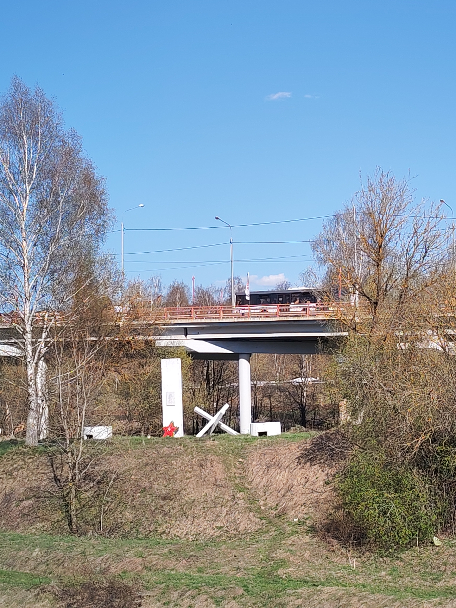 The image shows a memorial complex dedicated to the soldiers who died during the Great Patriotic War, located in Dmitrov, Russia. The complex includes a white concrete stele, a five-pointed red star, and stylized anti-tank obstacles. The memorial is situated on a grassy slope. In the background is a bridge with a bus crossing it. The trees are budding, suggesting spring. The sky is blue with few clouds. Street lamps are visible along the bridge.