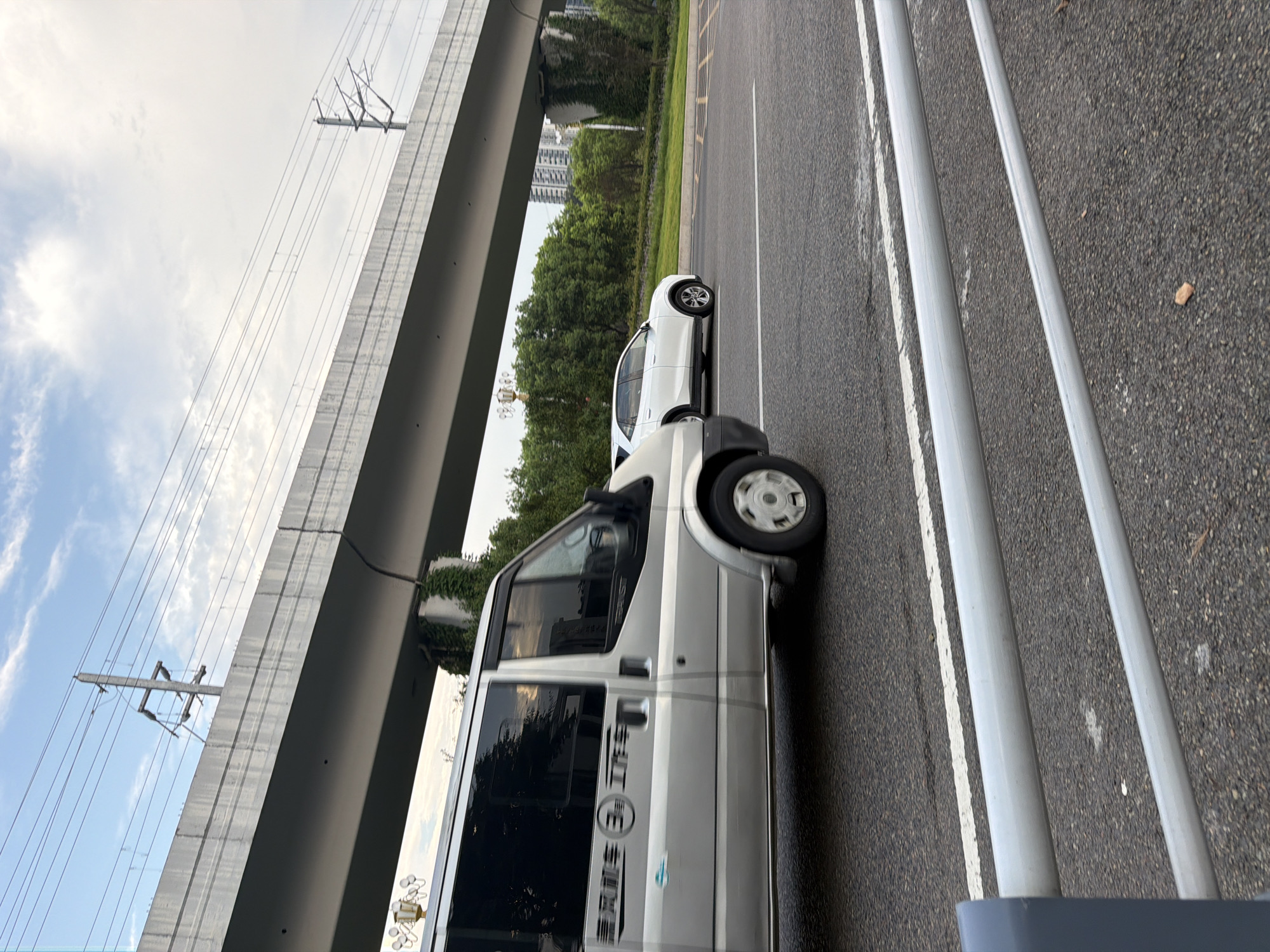 A silver van is traveling on an asphalt road beneath an elevated overpass. The overpass has a concrete structure with electrical lines running above it, supported by a utility pole. To the right of the van, a white SUV is also traveling in the same direction. The road has white lane markings and is bordered by a grassy median with trees. In the background, buildings and more trees are visible. The sky is partly cloudy. This scene is located in the city of Jiangjin, China, as indicated by the Chinese characters on the side of the silver van, which read "顺风车" (shùn fēng chē), meaning "ride-sharing car" or "free ride," and "三马车" (sān mǎ chē), meaning "three-wheeled vehicle," likely referring to a specific type of local transport.