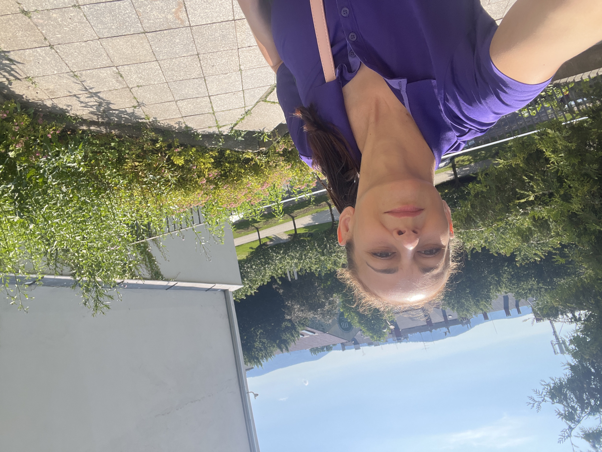 In a sunny, outdoor environment, a woman in a purple polo shirt is taking a selfie. She has light brown hair pulled back in a ponytail and is looking towards the camera with a neutral expression. The background shows a cityscape with buildings, trees, and rolling hills under a clear blue sky. The foreground consists of a tiled walkway and lush green bushes.