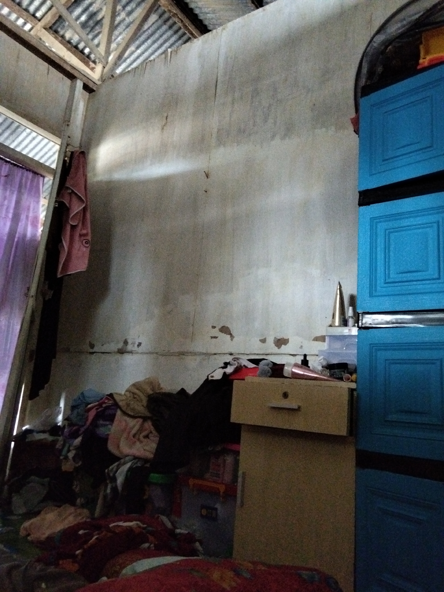 The image depicts a cluttered room interior. The walls are unfinished, showing patches of discoloration and damage. A corrugated metal roof is visible at the top, supported by wooden beams. To the left, a purple curtain hangs near an open doorway. A pink towel is draped over a wooden beam. In the foreground, a pile of clothing and fabrics is stacked on the floor. To the right, a tall blue cabinet with multiple drawers stands against the wall. In front of the cabinet, a small tan cabinet with a single drawer is placed. On top of the tan cabinet, several objects are visible, including a golden cone-shaped item, some bottles, and what appears to be a stack of clear plastic containers. The overall impression is of a storage area or a room with many items in disarray. The image is an interior shot and does not provide information to determine a specific location within Benuaraja, Indonesia.