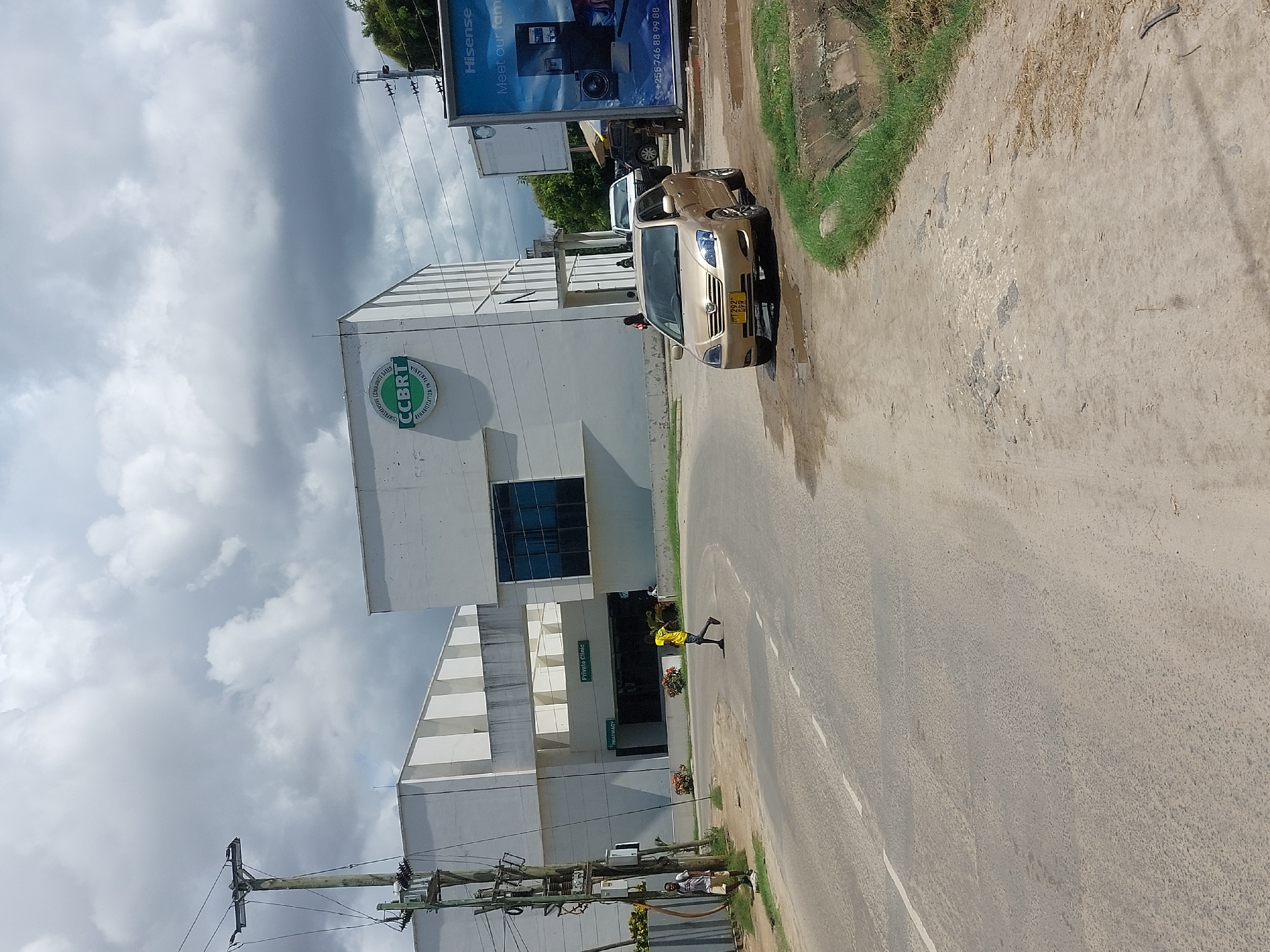 The image depicts a street scene in Dar es-Salaam, Tanzania, in front of the Comprehensive Community Based Rehabilitation in Tanzania (CCBRT) building, a healthcare facility. A gold-colored sedan is parked partially off the road. A person wearing a yellow shirt is seen crossing the street in front of the entrance to the "Private Clinic". A large Hisense billboard advertising consumer electronics is also visible. Utility poles with power lines are present on the left side of the image. The sky is overcast.