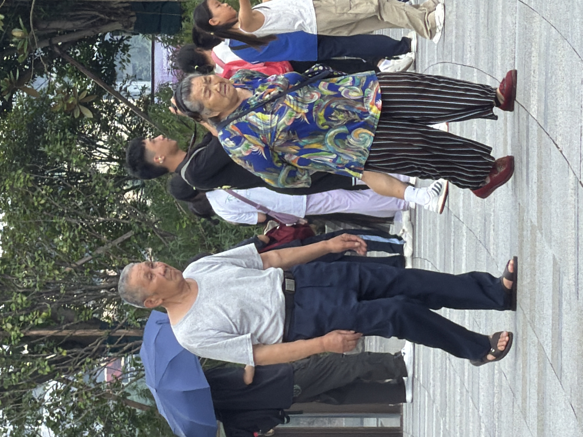 In the Yuzhong District of China, an elderly Asian man wearing a grey t-shirt and dark blue pants with sandals walks to the left. Beside him and slightly behind, an elderly Asian woman in a colorful, abstract patterned top and vertically striped pants with red slip-on shoes walks to the right. She appears to be carrying a dark blue crossbody bag. Behind them, a group of people are visible, including a young man with short dark hair wearing a black shirt, and several children. The background features trees and a paved walkway with a geometric pattern. A blue umbrella is partially visible to the left of the man.