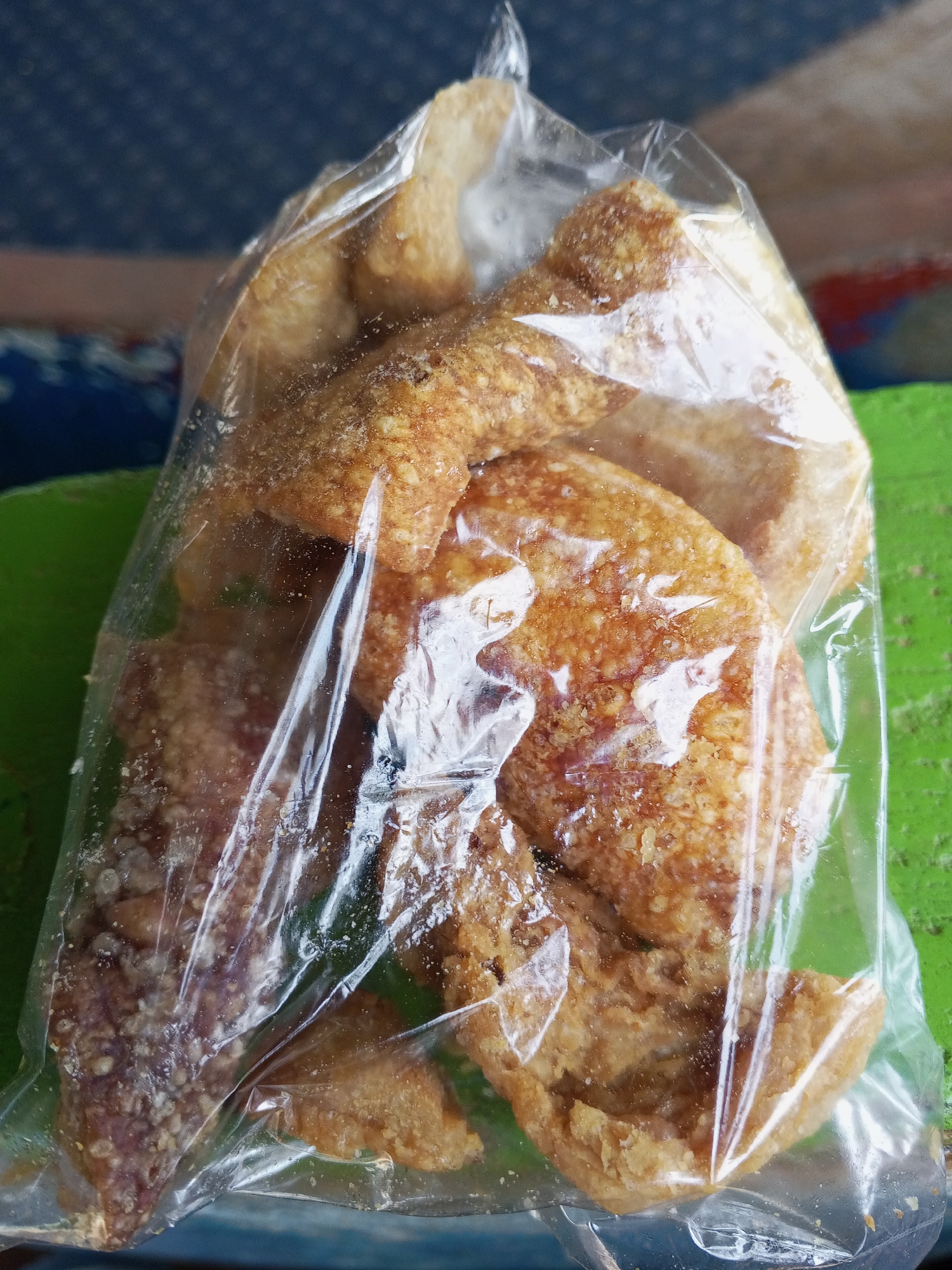 A plastic bag filled with fried pork rinds sits on a green and blue patterned surface. The pork rinds are irregularly shaped, golden-brown to dark brown, with a rough, crispy texture. The clear plastic bag is crumpled at the top, appearing closed but not completely sealed. The location is not specified beyond the generic "Unknown, Unknown."