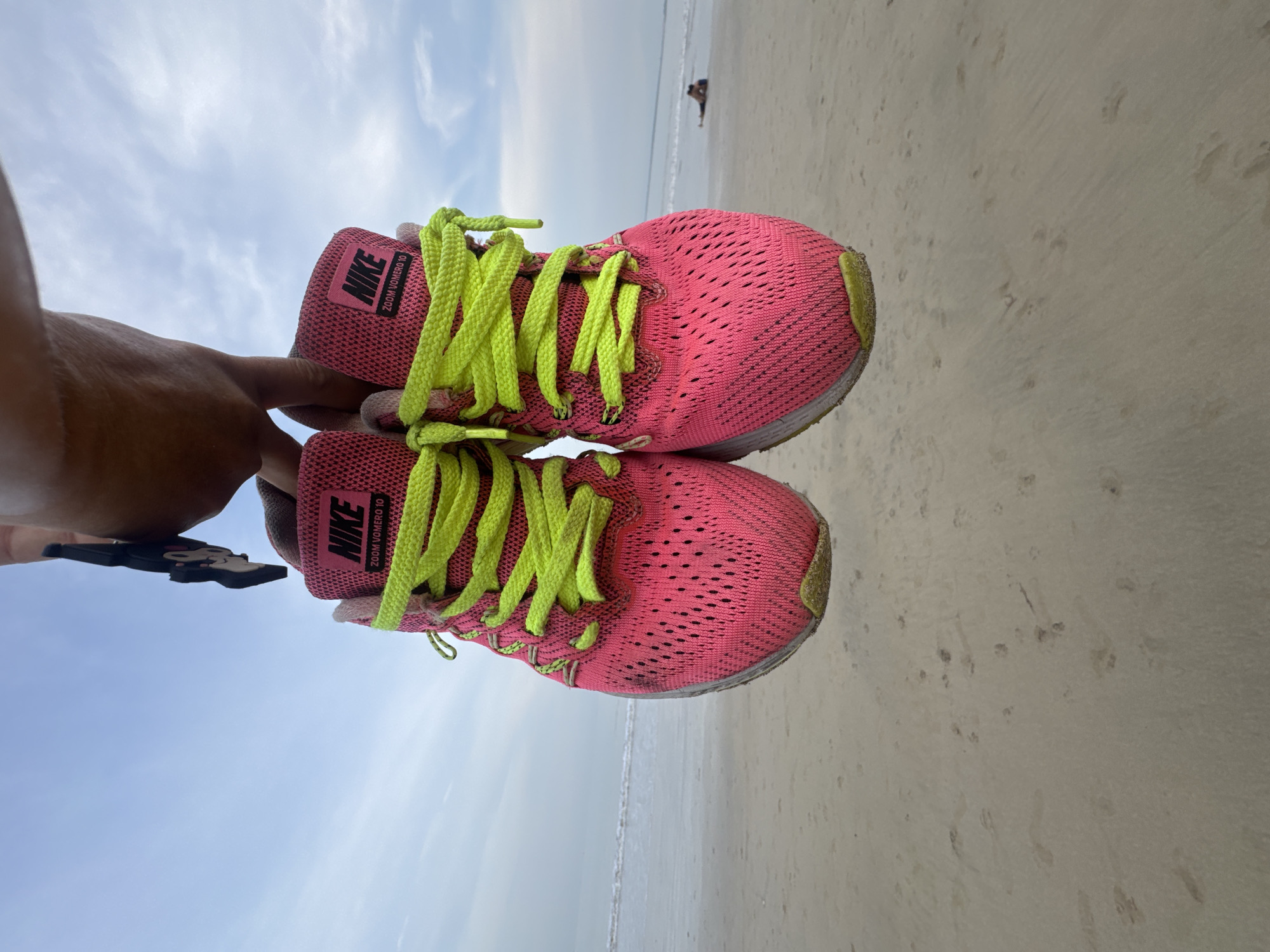 The image shows a pair of pink Nike Zoom Vomero 10 running shoes held up against a beach scene. The shoes have bright yellow-green laces and are slightly dirty, suggesting they have been used. The background features a sandy beach, likely in Jimbaran, Indonesia, with the ocean visible in the distance. The sky is partly cloudy. In the far background, there is a person on the beach near the water's edge. A portion of a person's arm and hand holding the shoes is visible on the left side of the image.