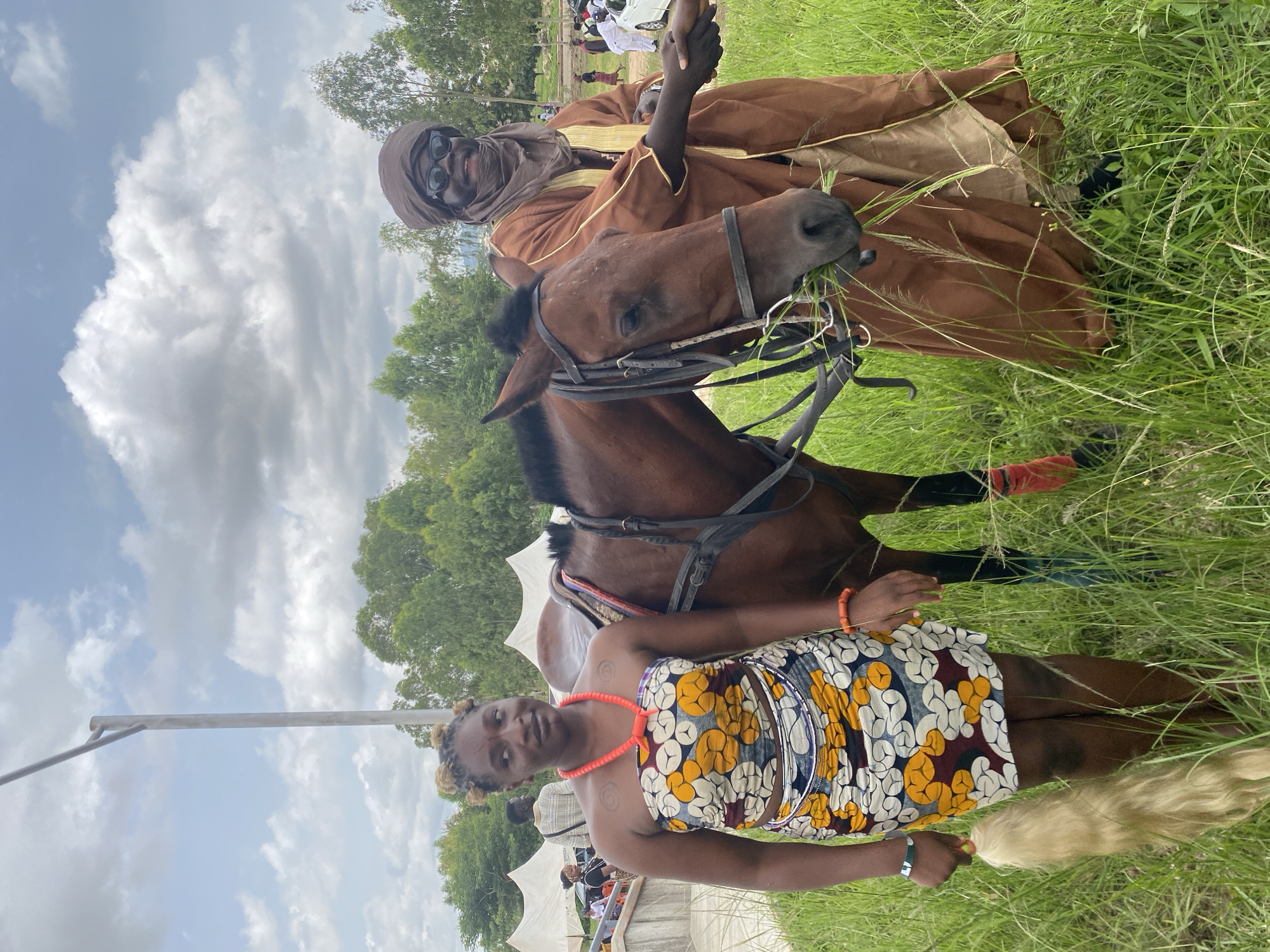 In Jos, Nigeria, a woman and a man stand beside a horse in a grassy field, under a cloudy sky. The woman, positioned on the left, wears a colorful strapless dress with a vibrant floral pattern and a thick red beaded necklace. She holds a bundle of light-colored hair in her left hand. The man, on the right, is dressed in a flowing brown robe and a headscarf, complemented by dark sunglasses. He is shaking hands with someone out of frame to his right. The horse, a brown equine with a black mane, is equipped with a bridle and appears to be eating grass. In the background, several white tents are visible, suggesting a potential event or gathering. Trees line the horizon, and a lamppost stands to the left of the frame.