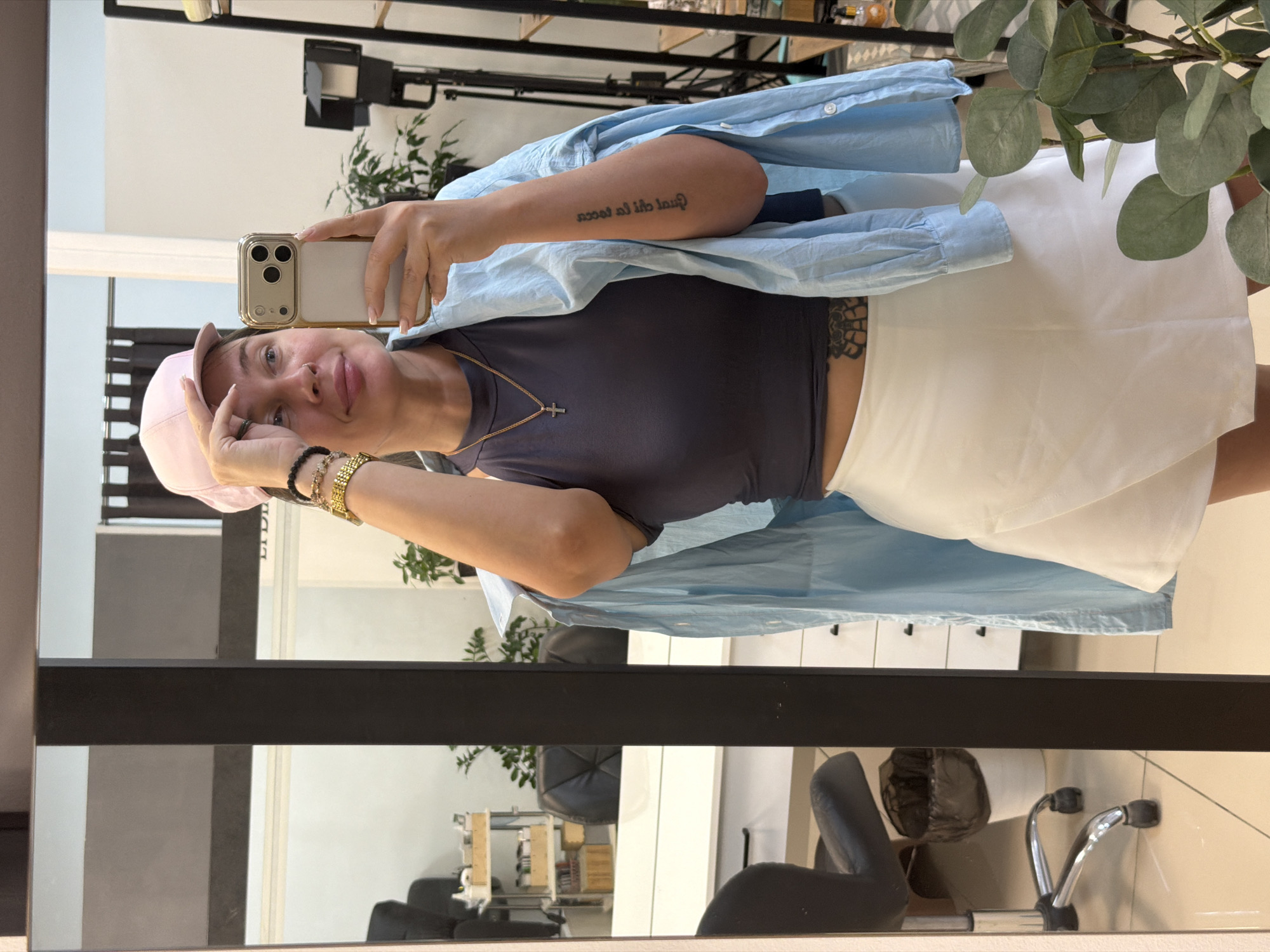 This media file features a mirror selfie of a young woman taken indoors, likely in a salon or studio in Chaikovsky, Russia. The image is oriented 90 degrees clockwise.
The woman, positioned in front of a large mirror with a black frame, is holding a gold-cased iPhone with three camera lenses to capture her reflection. She is wearing a light pink baseball cap, a dark gray or navy crop top, and a white skirt or skort. A light blue oversized button-up shirt is casually draped over her shoulders. Her accessories include a gold chain necklace with a silver cross pendant, multiple bracelets (one resembling a gold watch band, others beaded), and rings on her left hand. A tattoo with text in what appears to be a foreign language (possibly Italian), read backward in the reflection, is visible on her inner right forearm. She has a gentle smile or slight pout on her face, looking directly into the camera.
The background, visible through the mirror's reflection, depicts a modern interior space. Elements include shelving units, a plant with large green leaves (resembling eucalyptus), professional lighting equipment (such as a softbox or panel light), white cabinets, and black office or salon chairs. The setting suggests a well-lit, possibly commercial, environment. No specific time of day or weather details are discernible from the image, but the indoor lighting is bright.