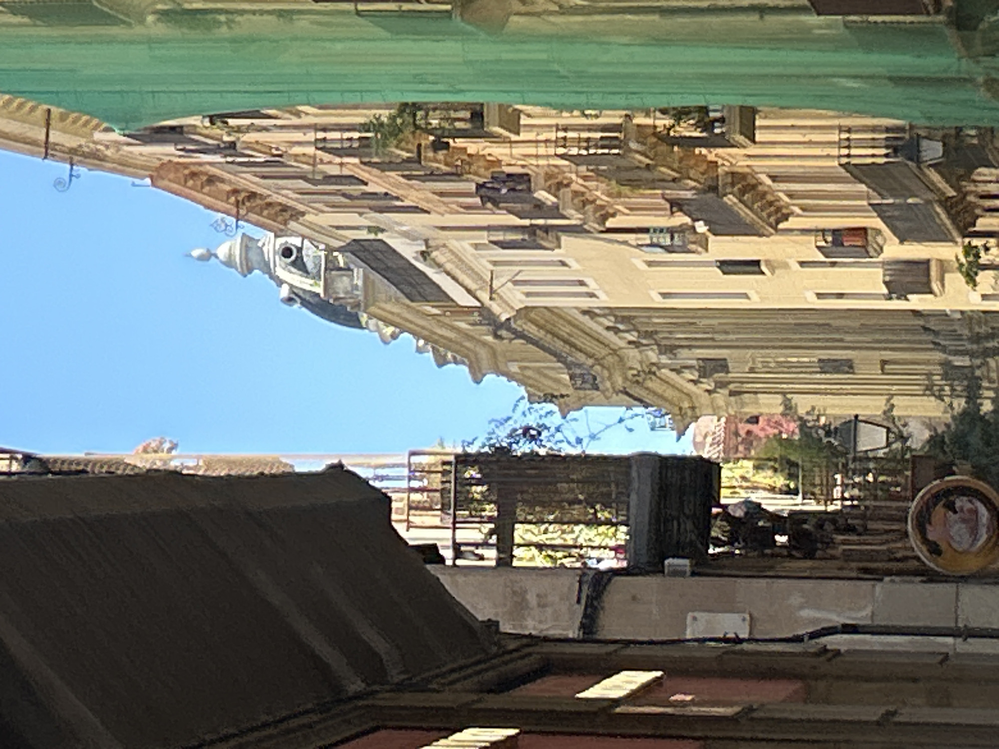 This image shows a view of buildings in Barcelona, Spain, seen from a lower rooftop perspective. In the foreground, a dark roof with brown tiles is visible. Behind this, a lighter-colored rooftop displays assorted objects including a dark rectangular box, a small brick pile, and a decorative plate. The background features a building with multiple floors, ornate architectural details and balconies. The sky is a clear blue and the image is inverted, which might reflect the light or create an unusual perspective.