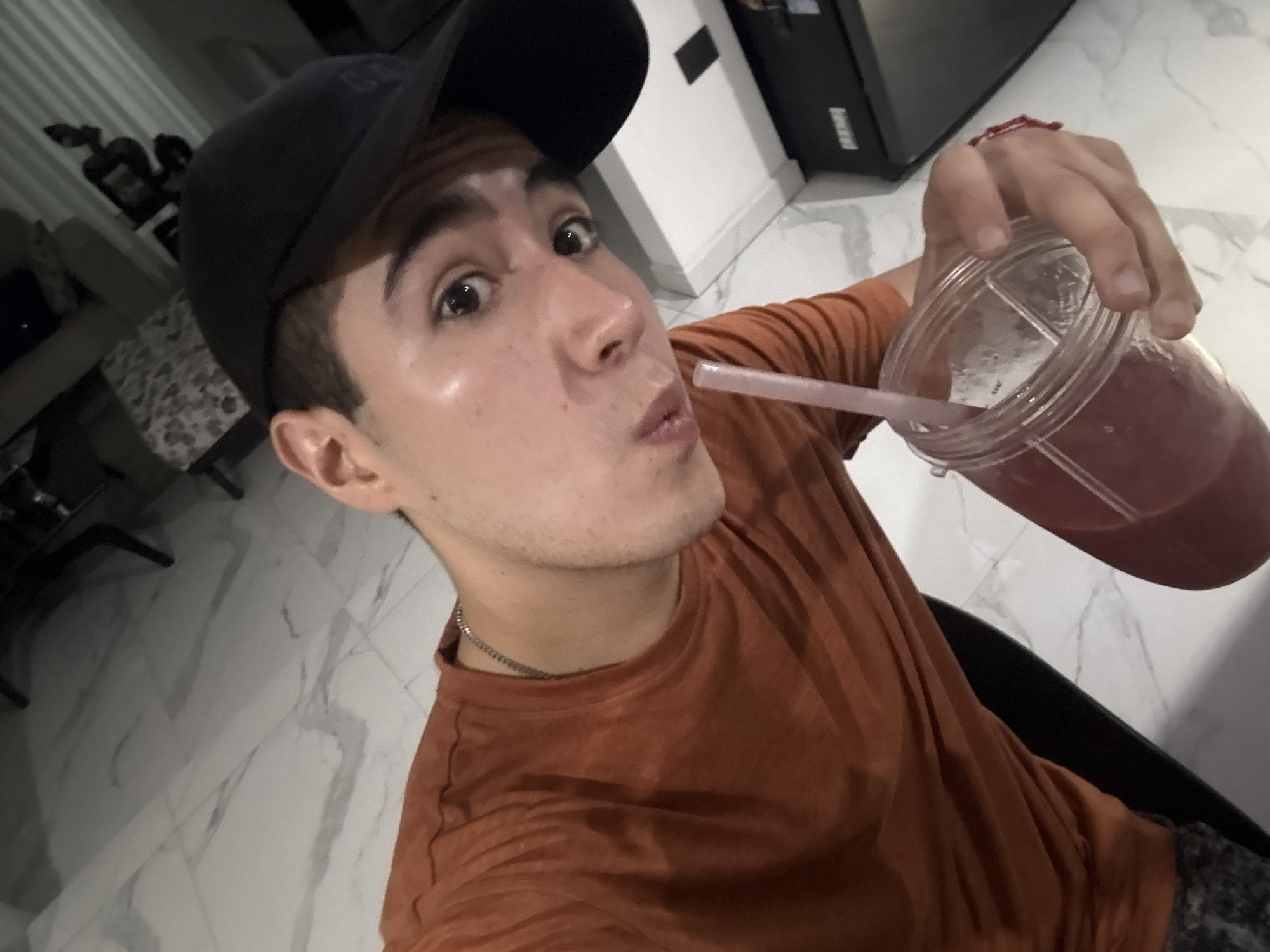 A young man wearing a black baseball cap and an orange t-shirt is holding a large plastic cup with a pink drink and a straw. He is pursing his lips as if drinking from the straw. He is wearing a thin silver chain. The background shows a marble-patterned floor, a white wall, and a glimpse of furniture. The scene appears to be indoors. There is no verifiable evidence to confirm the location as San Bernardino Tlaxcalancingo, Mexico.