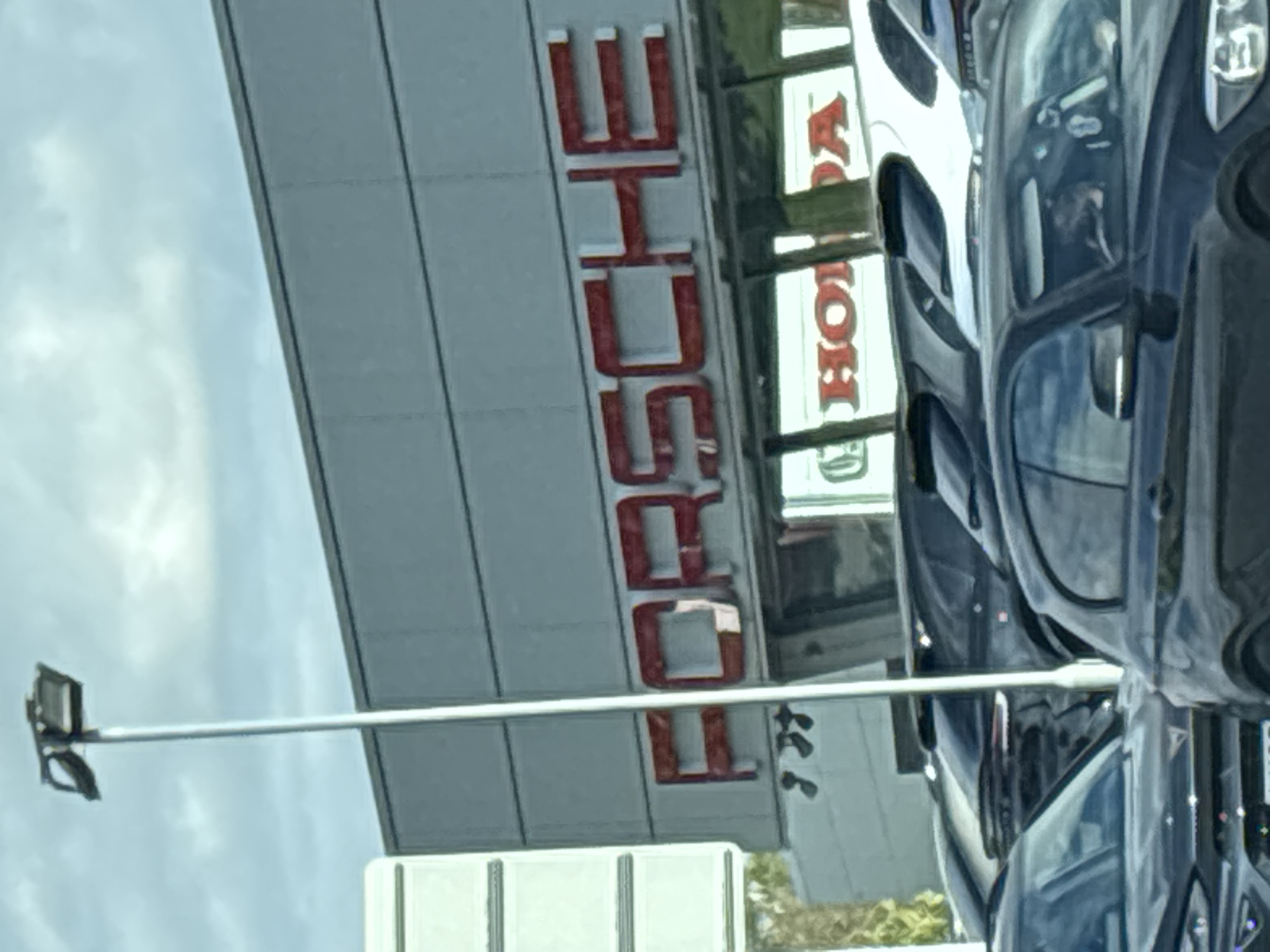 The image depicts the exterior of a building identified by signage as a Porsche dealership, located next to a Honda dealership. A row of cars is visible on the right. A streetlight is present in the foreground. The location is presumed to be Ellesmere Port, United Kingdom, based on the presence of these particular dealerships, but the exact location is not definitively confirmed by the image.