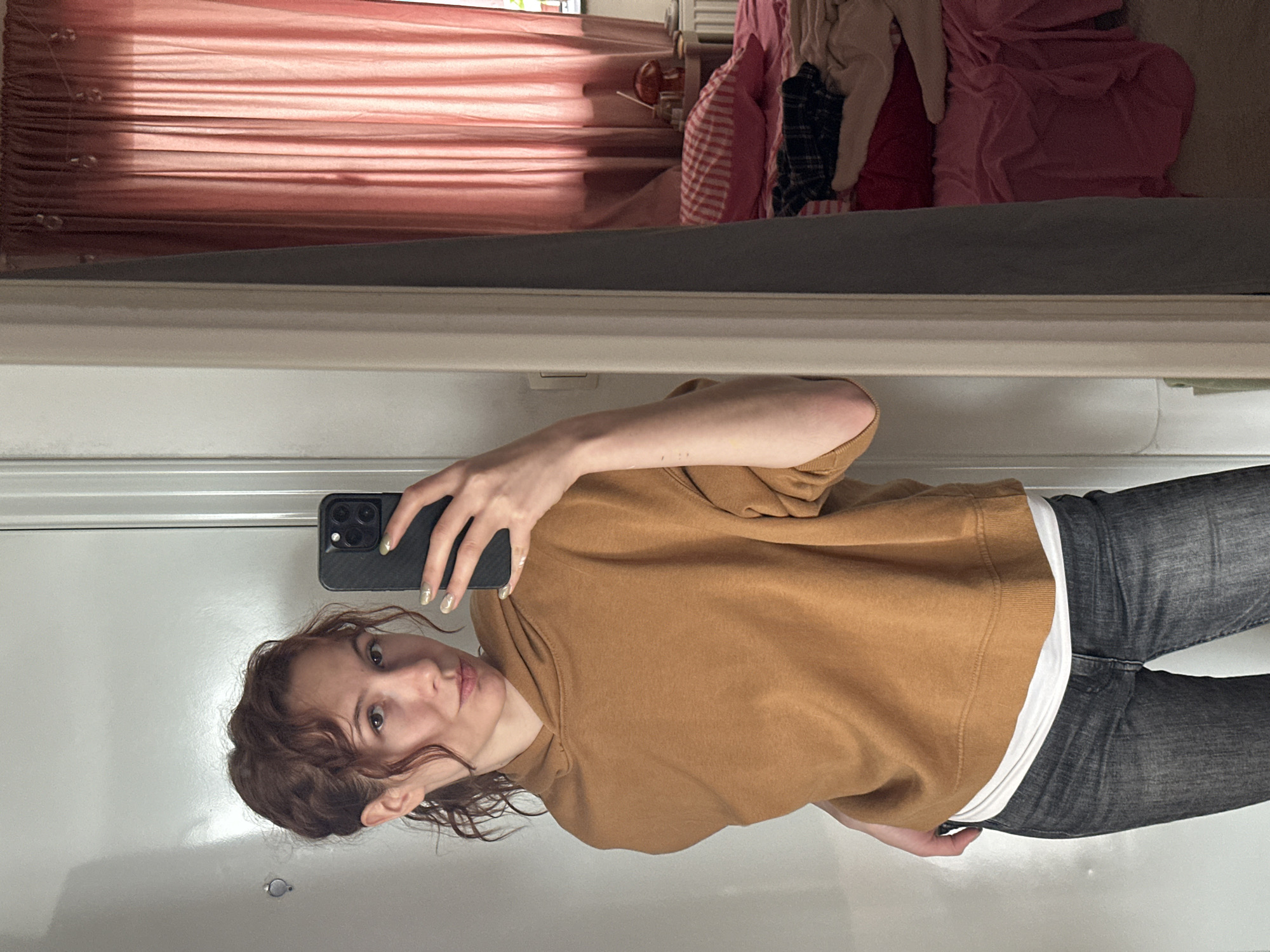 A person with curly auburn hair is taking a selfie in a mirror. The person is wearing a brown hoodie and grey jeans with a white t-shirt visible at the waist. The phone case on the phone held is black. The background of the mirror reflection shows pink curtains, a dresser with items on top of it, and clothes piled on top of the dresser. The location is unknown and cannot be determined based on this image.