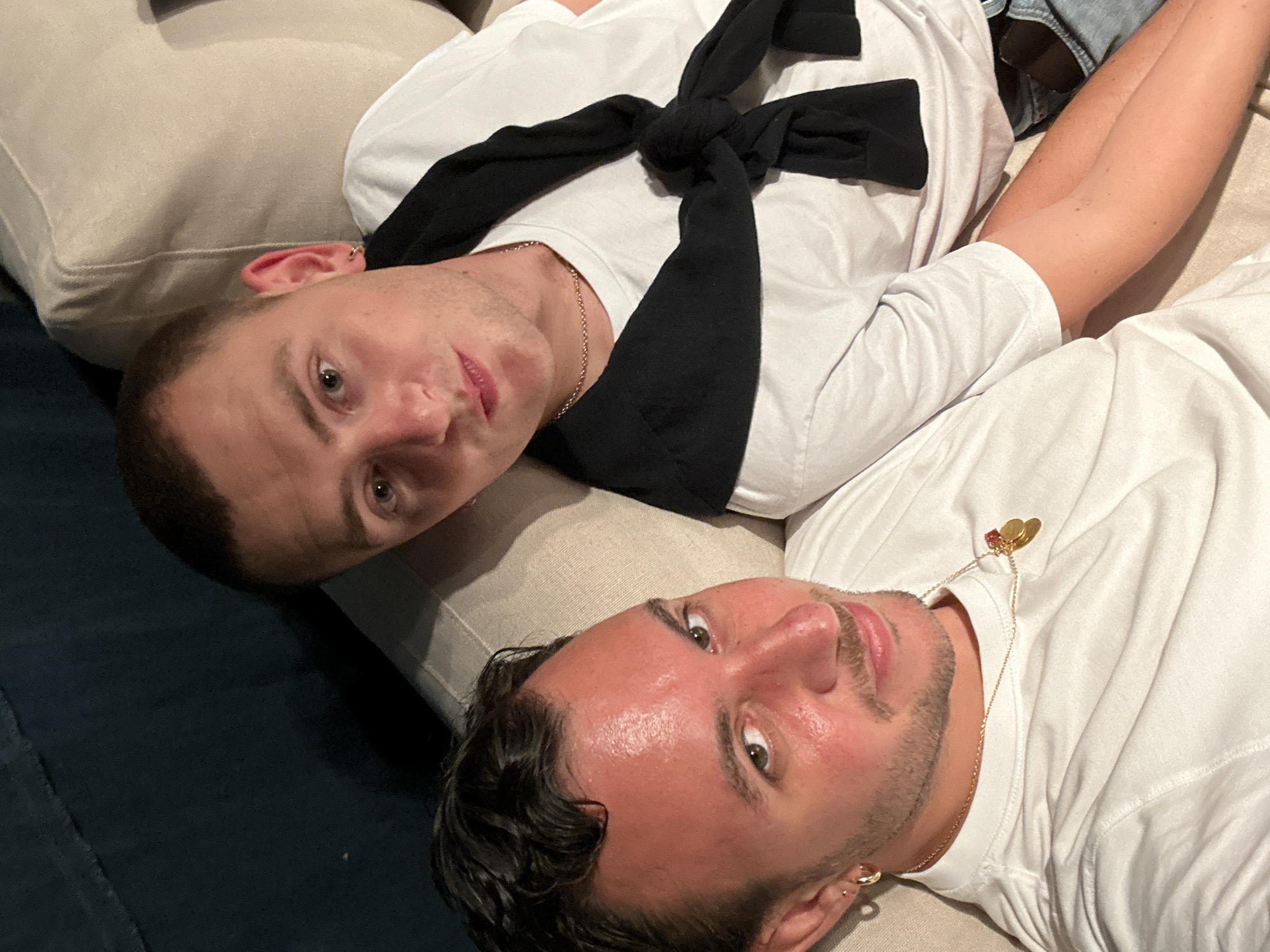 Two young men are photographed from a high angle, reclining on a light-colored couch. The man in the foreground, on the left, is facing forward and smiling slightly. He has dark, curly hair and is wearing a white t-shirt and gold jewelry, including hoop earrings and a necklace with charms. His skin appears flushed. The second man, on the right, is partially visible and angled towards the camera. He has short, light brown hair and is wearing a white t-shirt with a black sweater tied around his neck. Both men are casually dressed. The background includes a dark blue surface, possibly a sofa or wall, and the textured beige upholstery of the couch they are seated on. The lighting suggests an indoor setting, likely a home. The image does not contain any specific identifiable landmarks or indicators of its location within London, United Kingdom.