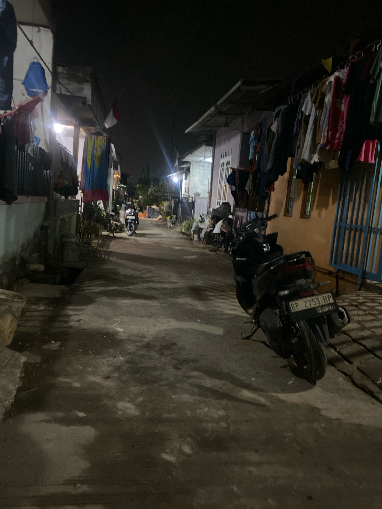 An outdoor, low-light, eye-level shot of a narrow alleyway at night. The alley is paved with concrete and has a damp, patchy appearance. On the left side of the alley, there is a light-colored building with laundry hanging on a clothesline in front of it. A blue bag and red clothing are visible hanging there. Further down the alley on the left, an Indonesian flag is displayed. On the right side, there is an orange-colored building with a blue metal gate and laundry hanging overhead. A black motorcycle with a license plate reading "BP 2753 RP" is parked in the foreground on the right side of the alley. Another motorcycle is visible further down the alley on the left. The scene suggests a residential area with limited lighting.