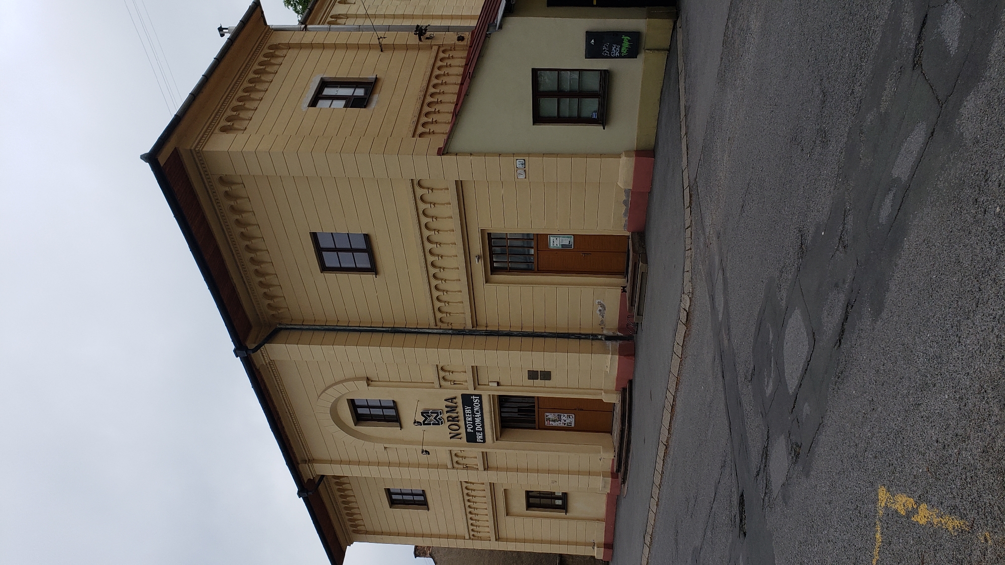 The image shows the exterior of the NORMA store, a business located in Prešov, Slovakia. The building is two stories high and painted a light yellow color, with decorative elements along the roofline and around the windows. The store entrance is centrally located on the first floor, marked by a sign with "NORMA" in large letters. Below this, it says "POTREBY PRE DOMÁCNOSŤ", which translates to "Household Needs". There are two sets of double wooden doors, the left set being the main entrance. Smaller windows are visible on both floors. To the right of the main building, a smaller, single-story addition with a red roof is attached. This addition has a dark-framed window and a sign on the wall. The street in front of the building is paved with asphalt and shows signs of wear, with some cracks and patches. The sky is overcast.