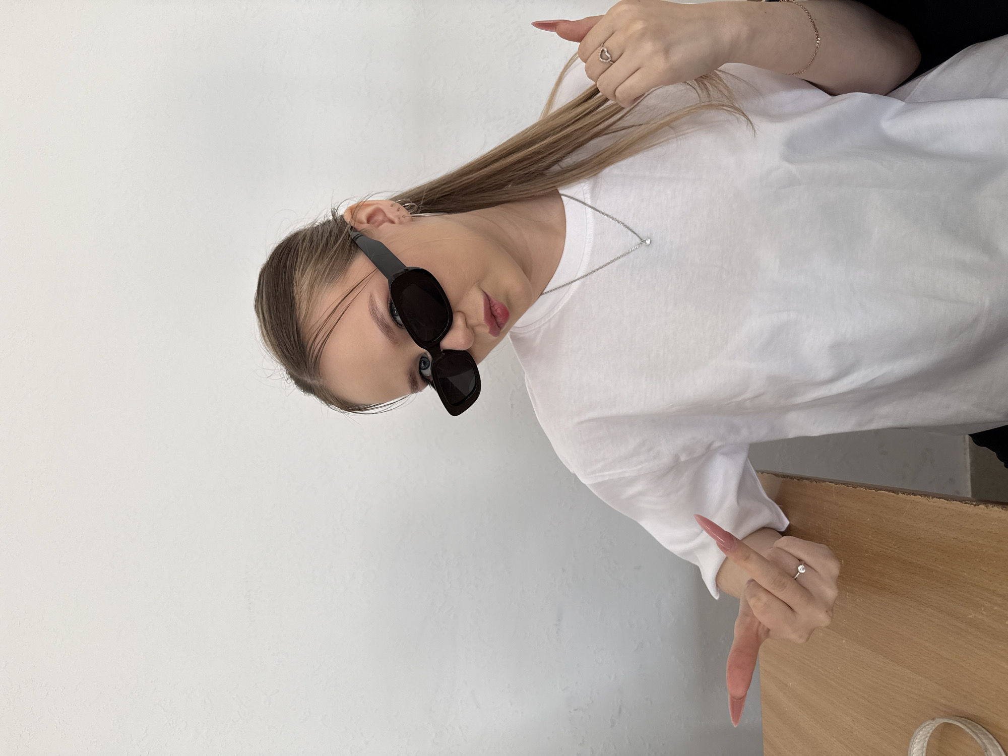 A young woman with blonde hair wearing black sunglasses and a white t-shirt is positioned diagonally in the frame. She is wearing a silver necklace and jewelry on her fingers. Her long, pink nails are noticeable. The background consists of a white wall on the left and a wooden surface on the right. The location is Chaikovsky, Russia.