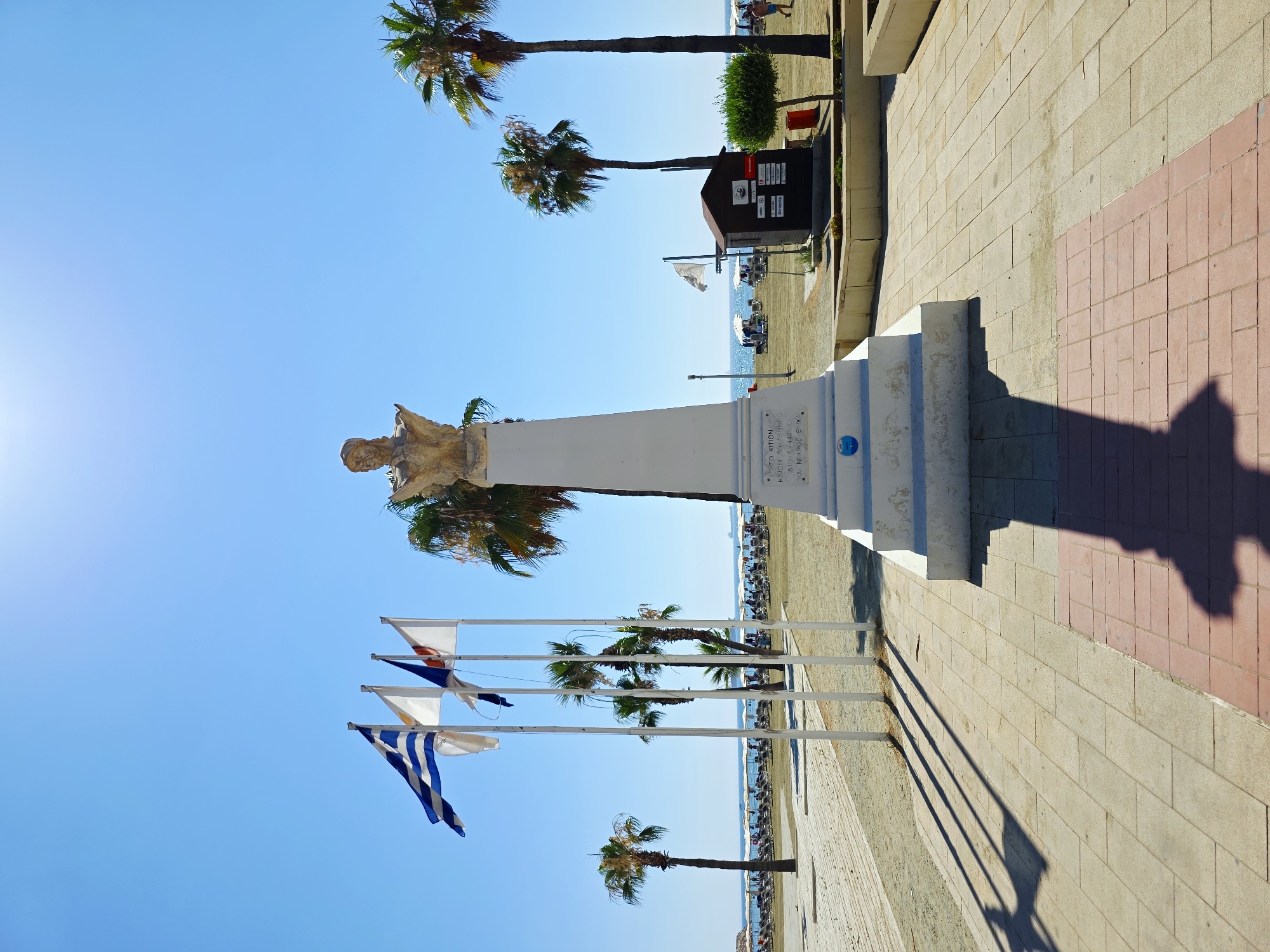This is a vertical, eye-level, full-shot photograph taken on a bright, sunny day in Larnaca, Cyprus. The foreground is paved with light-colored square tiles, with a section of red tiles on the right. A large, white monument with a bust of a man on top stands prominently in the center, casting a long shadow. To the left of the monument, three flagpoles fly flags, including the flag of Greece. Palm trees are scattered throughout the scene, with one on the far left and two taller ones behind the monument. In the middle ground, a sandy beach is visible, lined with numerous lounge chairs and umbrellas. The sea is calm and blue in the background. To the right of the monument, a small, dark-colored wooden structure with signs is visible, and a person in blue shorts walks past it. The sky is a clear, vibrant blue with a bright sun at the top center, creating a lens flare. The overall scene depicts a typical beachside promenade in Larnaca, likely near the Finikoudes promenade, a popular tourist area known for its palm-lined street, monument, and beach.