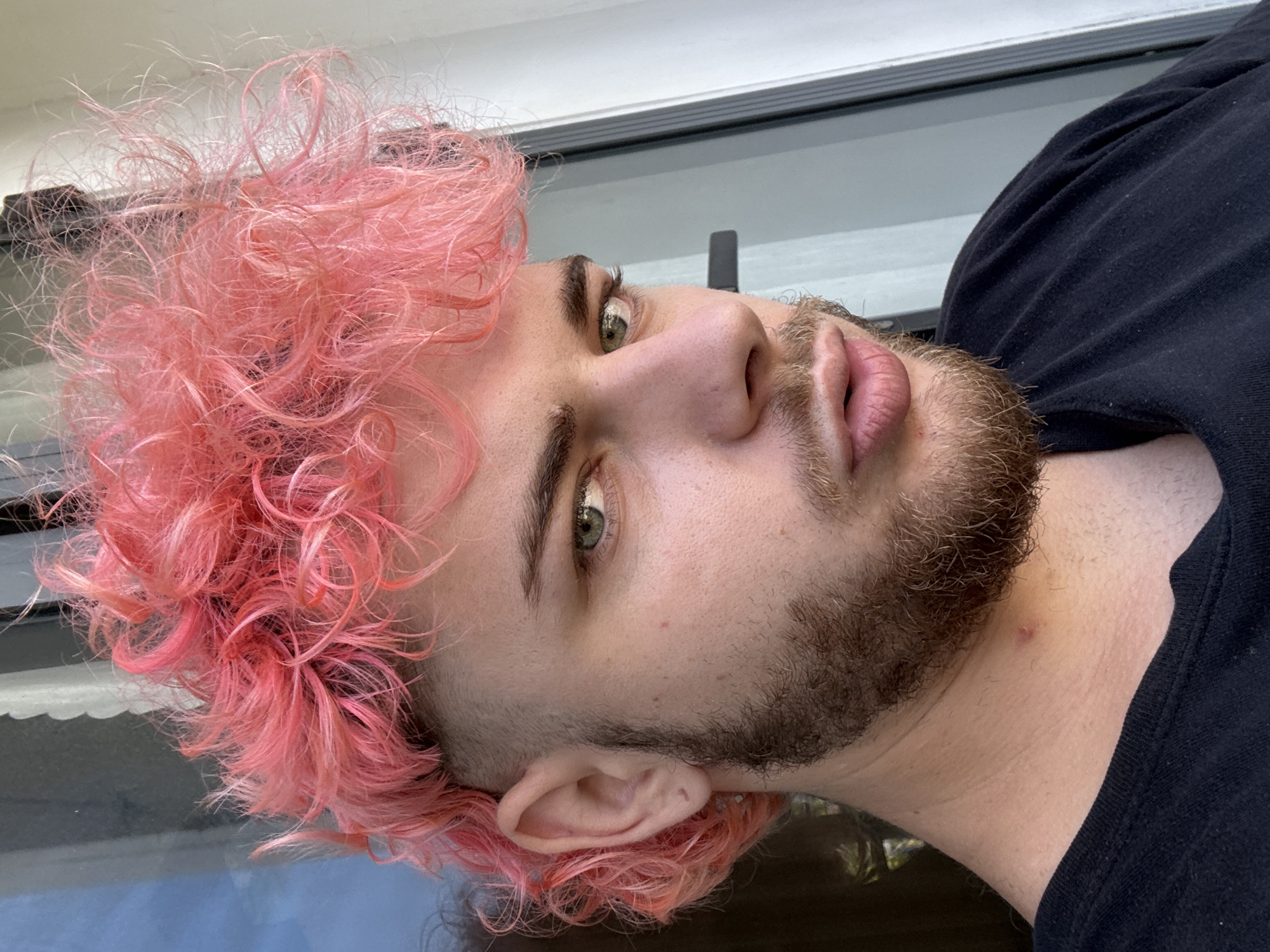 A close-up headshot of a young man with pink curly hair and a beard. He has light skin and green eyes. He is wearing a black shirt. The background appears to be outdoors with a glimpse of a building or window frame visible. There is no evidence to suggest this image was taken in Đà Nẵng, Vietnam.