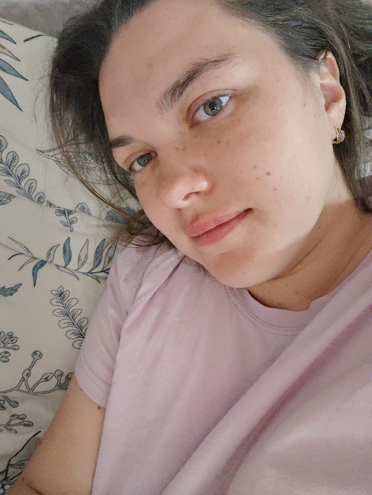 A woman is lying on a bed with floral patterned sheets. She is wearing a light purple t-shirt. Her face is visible and she has light brown hair, fair skin, and green eyes. She has some freckles on her face. She is wearing earrings. The location is not identifiable as being in the city of Chaikovsky, Russia, from the details visible in the image.