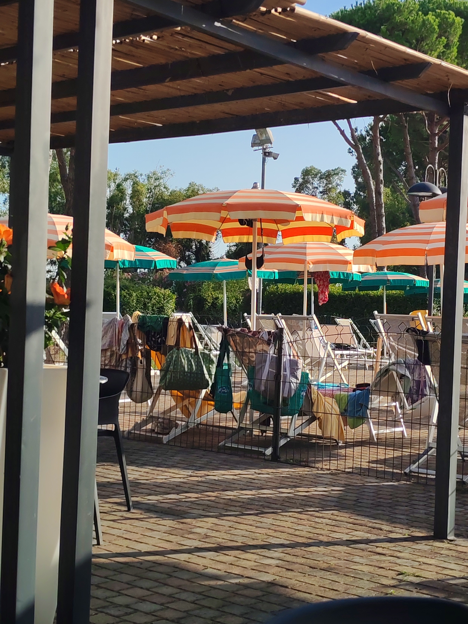 The image depicts an outdoor scene under a wooden pergola, likely at a beach resort or public pool area in Luni, Italy. The foreground is partially obscured by vertical gray beams, suggesting the viewpoint is from under a covered structure. Beyond these beams, numerous beach chairs are visible, some occupied and some empty.
A collection of beach umbrellas, predominantly in orange and white stripes, are set up, providing shade. Several smaller, green umbrellas are also present. Hanging on a wire mesh fence in front of the chairs are various bags and towels, indicating recent use of the area. These items include at least two large green tote bags, a patterned bag, and several towels in different colors and textures. One chair in the background has a towel draped over its back.
The ground is paved with light-colored, square bricks arranged in a grid pattern, casting distinct shadows from the pergola and other structures. Lush green foliage and trees form the background, with a clear blue sky visible above. The overall impression is one of a relaxed, sunny day at a recreational facility. There are no people clearly visible in the image.