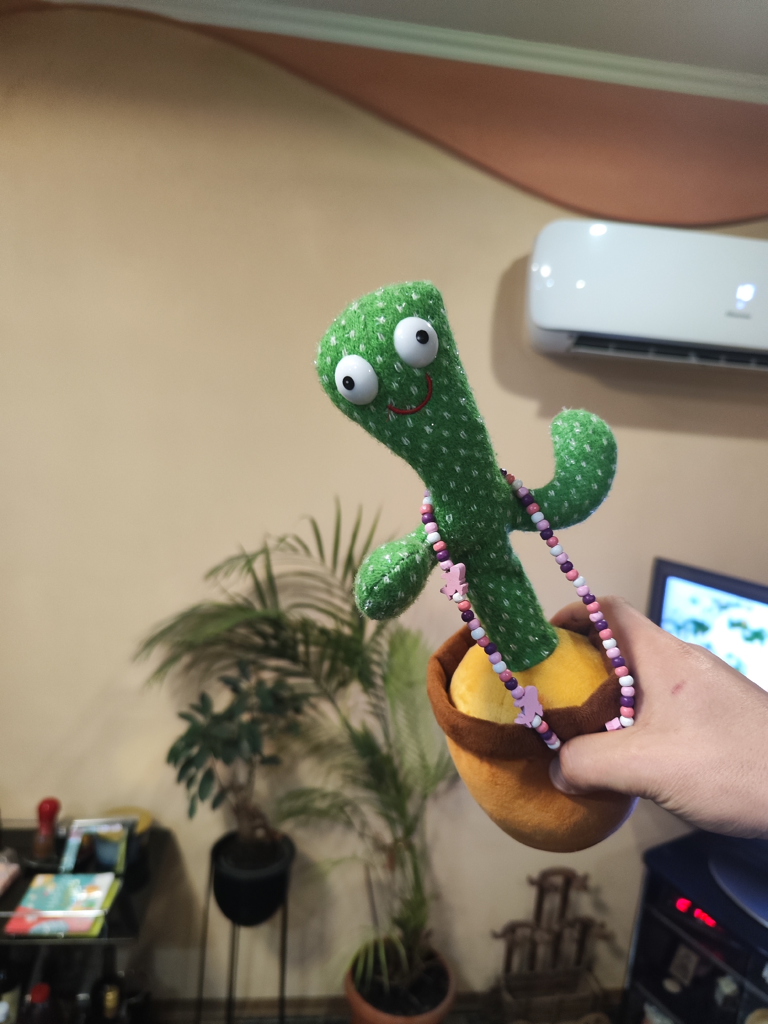 A plush green cactus toy wearing a pink and purple beaded necklace is being held in front of a tan wall in an indoor setting. The cactus is in a plush yellow pot with a brown rim. An air conditioning unit is mounted on the wall in the background, above a television. Two potted plants stand near a black table, and a decorative statue is visible in the background. The image appears to have been taken in a domestic setting. It is not possible to determine the location as Burgas, Bulgaria, from this image alone.