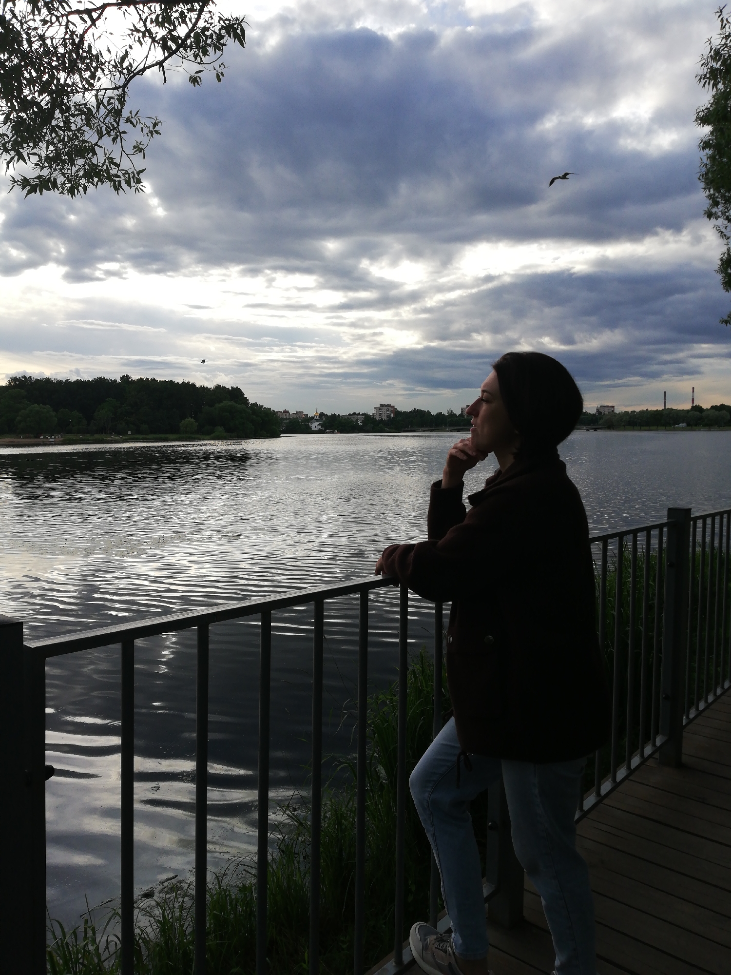 A woman stands on a wooden boardwalk, leaning on a metal railing that overlooks a large body of water. She is looking out towards the horizon, with her chin resting on her hand, appearing to be in deep thought. She is wearing a dark, possibly brown, jacket and light blue jeans. Her hair is short and dark, and she has a dark beanie on her head. The sky is overcast with dramatic, grey clouds, and shafts of light break through in places. A bird is visible flying in the distance, against the clouds. The water is rippled, reflecting the sky and the dark green trees that line the opposite bank of the lake or river. In the distance, beyond the trees, are some buildings and what appear to be industrial chimneys. The time of day appears to be late afternoon or early evening, with the diffused light suggesting an overcast sky. The overall mood is one of quiet contemplation and a connection with nature. The scene is set in Kolpino, Russia.