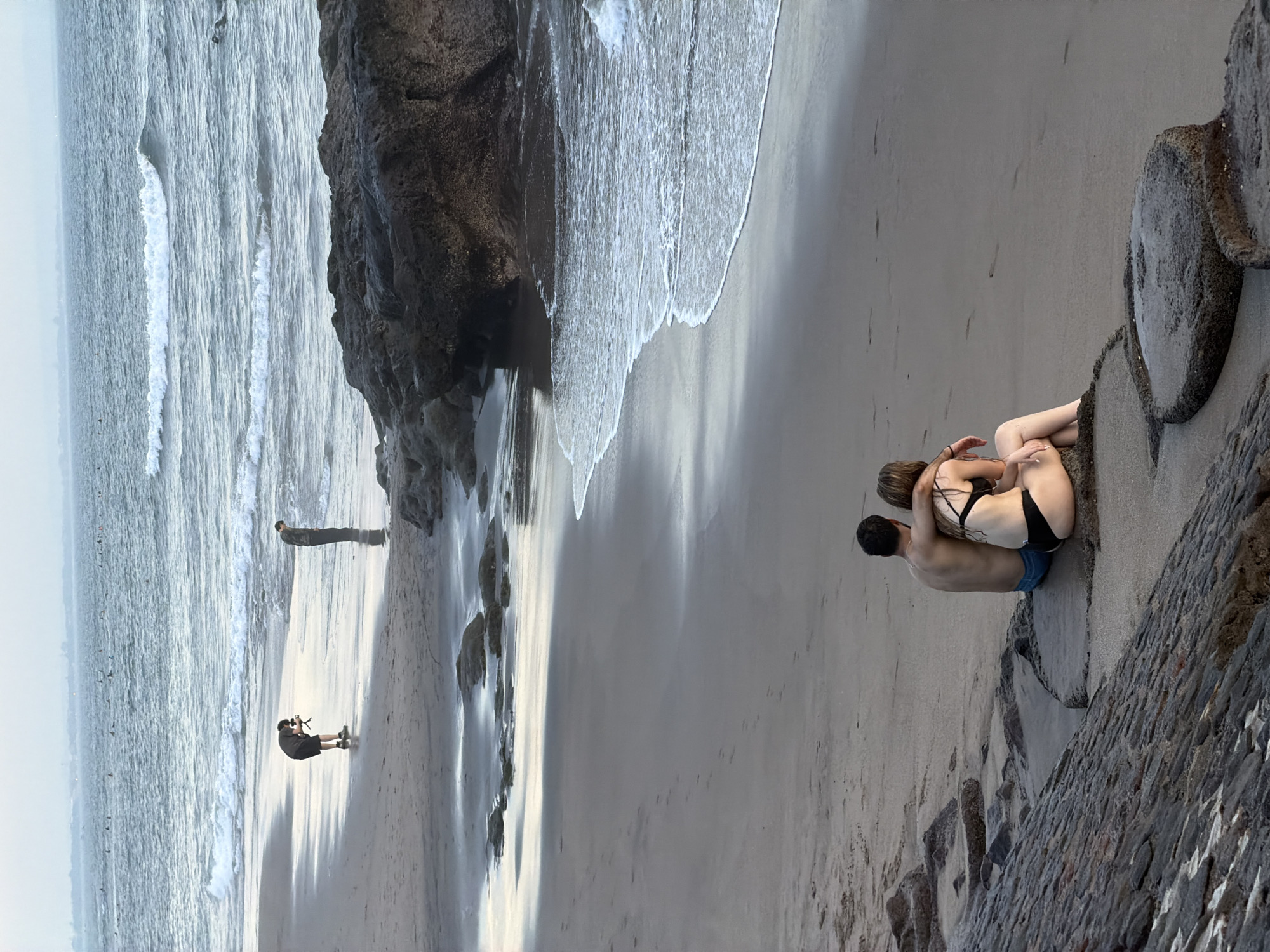 Here's a detailed description of the image:
**Location:** The image appears to be taken at a beach in North Kuta, Indonesia.
**Scene:** The scene depicts a beach environment with the ocean, sand, and rock formations.
**People:**
* A couple is embracing on the beach near a rock or wall structure. The woman is wearing a black bikini. The man is shirtless and wearing blue shorts.
* There are two other individuals on the beach in the distance. One appears to be standing, and the other is kneeling with a camera.
**Objects:**
* Large rock formations or cliffs are present along the shoreline.
* The beach has light-colored sand.
* Ocean waves are visible, indicating an active shoreline.
* There is a rock or wall structure near the couple, possibly a man-made structure.
**Actions/Interactions:**
* The couple is embracing, suggesting a close relationship.
* The person with the camera is likely taking pictures of the beach and possibly the couple.
**Environment:**
* The overall environment is a beach scene with a blend of natural elements (ocean, rocks, sand) and potential man-made structures.
* The lighting appears soft, indicating it might be early morning or late afternoon.
