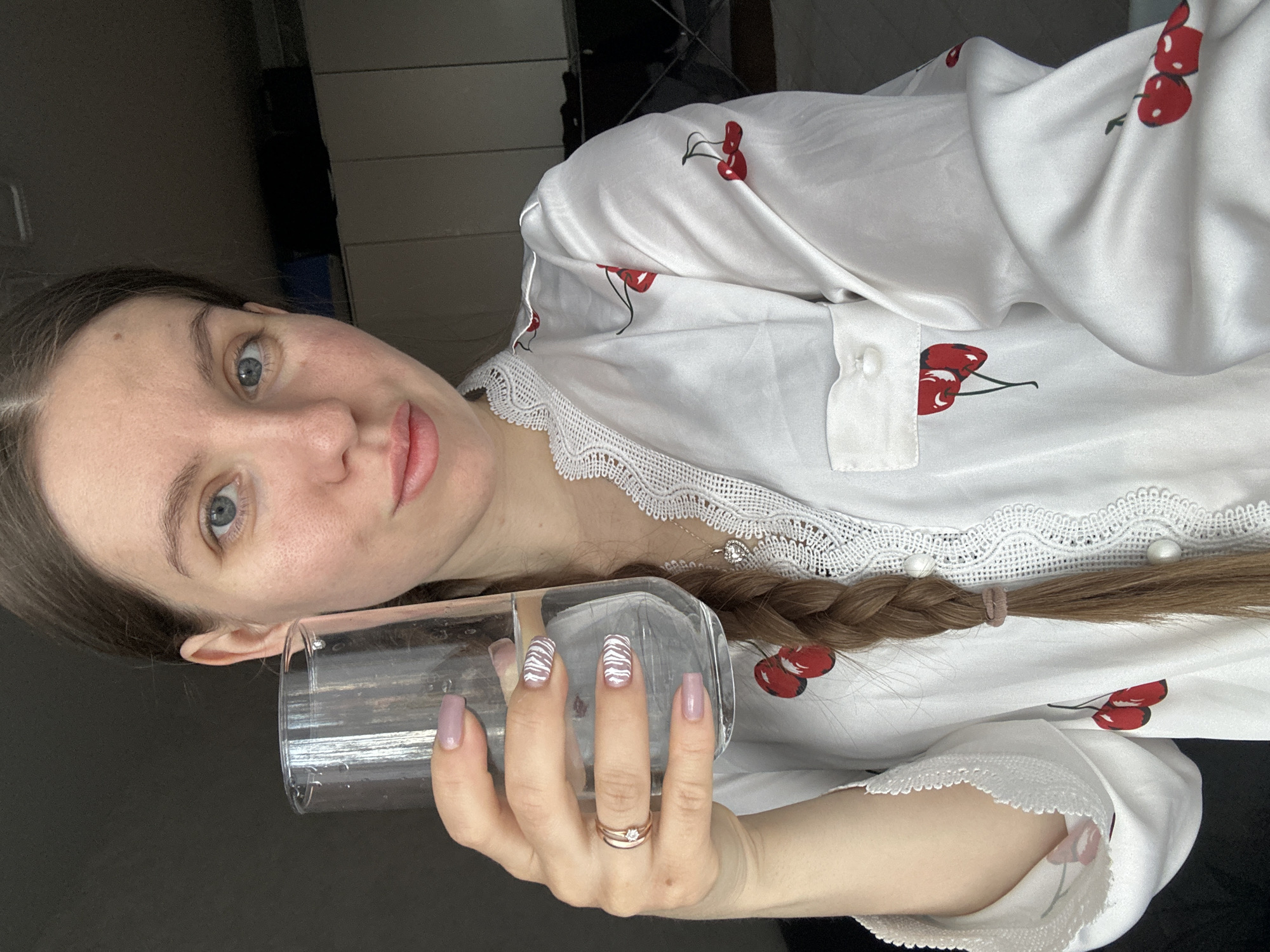 A light-skinned woman with blue eyes and brown hair braided to her right side is holding a clear glass in her right hand. She is wearing a white pajama top with a cherry print and lace trim. A ring is visible on her finger. The background is dark, suggesting an indoor setting. There is no information to support the location being Klin, Russia.