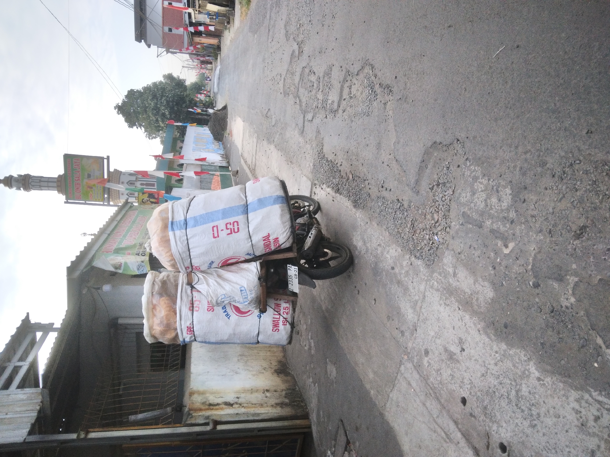 A motorcycle loaded with two large sacks of goods is parked on a paved road in Desa Pamijen, Kecamatan Sokaraja, Indonesia. The sacks are white with blue stripes and have writing on them, including "SWALLOW S ISI 25 L" and "SINDAL NSIN". The motorcycle has a license plate that reads "Z 2335 PA CB-31". In the background, there is a mosque with a minaret, a building with a sign that says "BU ONIH SALA JAYA", and several houses with red and white flags lining the street. The road is in poor condition with many potholes. The sky is overcast.