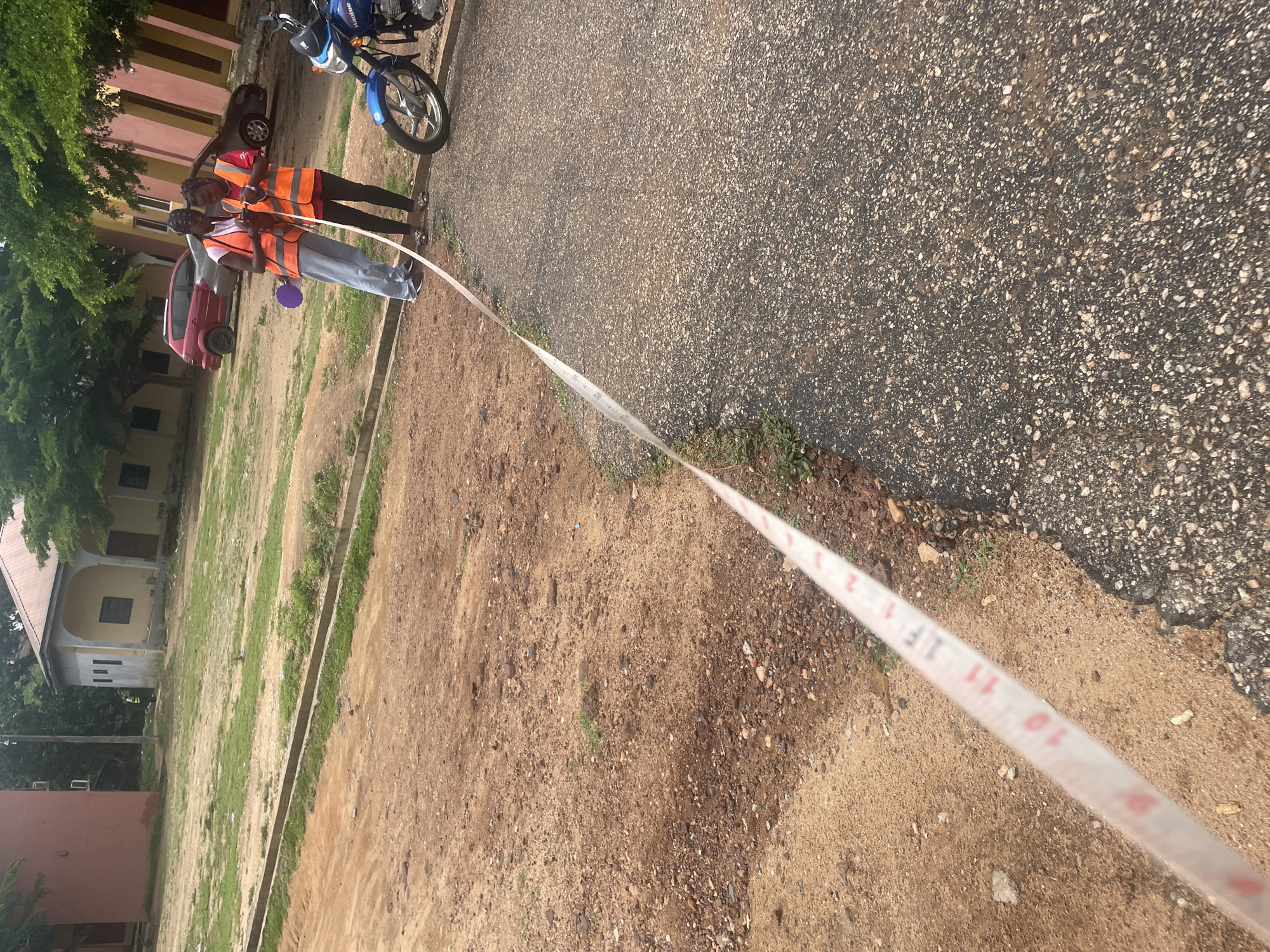 In Lokoja, Nigeria, two individuals wearing orange safety vests are standing on a paved road next to a dirt path, holding a white measuring tape. The tape is extended diagonally across the foreground, displaying red numerical markings from 9 to 11. Behind them, a blue motorcycle is parked on the right side of the road. Further in the background, several buildings with different colored facades are visible, along with parked cars and trees. The ground is a mix of dirt, gravel, and patches of grass. The overall scene suggests an outdoor measurement or surveying activity in a semi-urban or educational setting.