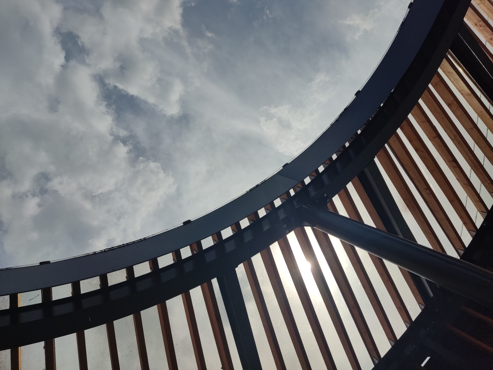 The image shows the underside of a curved, modern roof structure. The roof is composed of parallel wooden beams supported by dark metal framing and a large, dark, curved beam that forms the main arc. The sky, filled with white and gray clouds, is visible through the gaps between the wooden beams, and the sun is partially visible, creating a lens flare. A thick, dark cylindrical support column is visible in the lower part of the image, angled upwards. The perspective is from below, looking up at the sky through the architectural elements. The image does not contain any people or identifiable landmarks that would suggest a specific location in Noginsk, Russia.