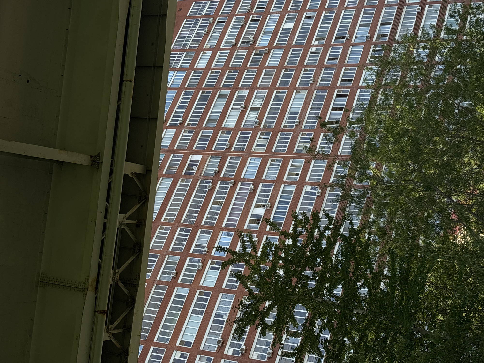 The image presents a view of a tall, red brick building with multiple rows of windows, each fitted with a white frame and an air conditioning unit. A green, structural element of a bridge or elevated structure is positioned in the foreground on the left, while foliage from trees obscures parts of the building on the right. The location is indeterminate.