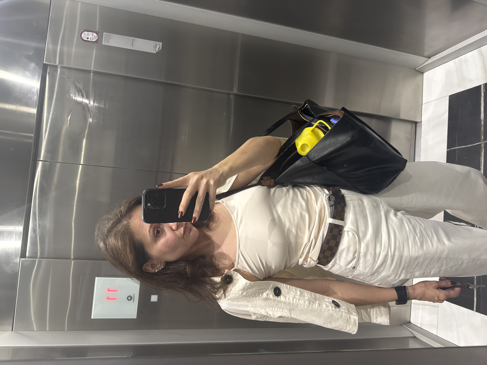 A woman is taking a selfie in an elevator. The elevator has stainless steel walls and a control panel that shows the number "11." She is wearing a cream-colored top, white pants, and a brown belt. She also has a black shoulder bag with a yellow object inside. The floor of the elevator is tiled in black and white. The location is most likely Dubai, United Arab Emirates, but this cannot be confirmed from the provided image alone.
