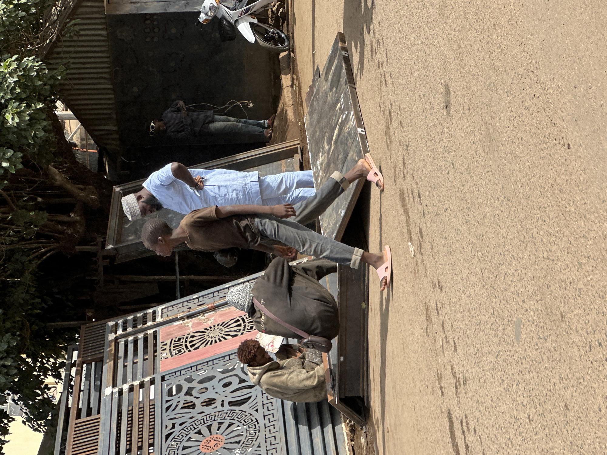 The image depicts an outdoor scene on a street in Jos, Nigeria, possibly a market or workshop area. In the foreground, a young boy in a brown t-shirt and grey pants walks away from the camera, wearing pink sandals. To the left, two men are seated on metal structures. One man wears a tan hooded sweatshirt, and the other, wearing a grey knitted cap, has a dark jacket and a bag slung over his shoulder. Behind them, large decorative metal gates are stacked. Further back, a man in a light blue traditional outfit and a white cap stands near a building with corrugated metal siding. Another person in dark clothing and sunglasses is visible further to the right, near a white motorcycle. The ground is paved with asphalt, and sparse vegetation is visible above the structures. The overall atmosphere suggests a commercial or industrial area.