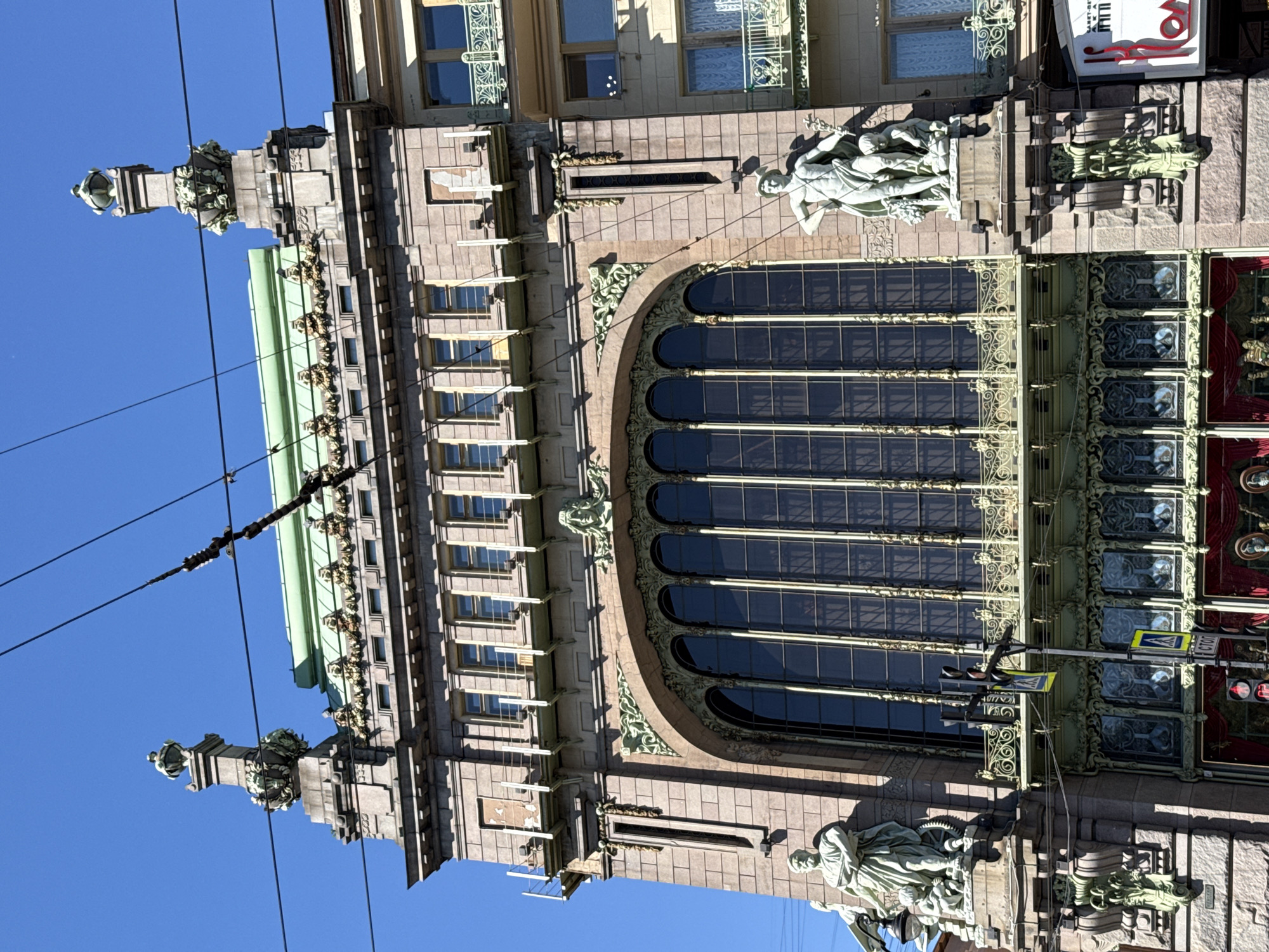 The image shows the Singer House, also known as the Dom Knigi, located on Nevsky Prospekt in Saint Petersburg, Russia. It is a prominent Art Nouveau building with a distinctive glass dome. The facade is adorned with architectural sculptures and decorative elements. In the foreground, there are power lines crisscrossing the sky, which is clear and blue. The building features large arched windows with intricate metalwork, and statues are placed on pedestals on either side of the main facade. Below the large windows, there are smaller, decorative windows. The street level appears to have shops with red curtains visible. There are traffic lights and street signs in front of the building. The architecture suggests a sunny day with clear visibility. No people are clearly discernible in the image.