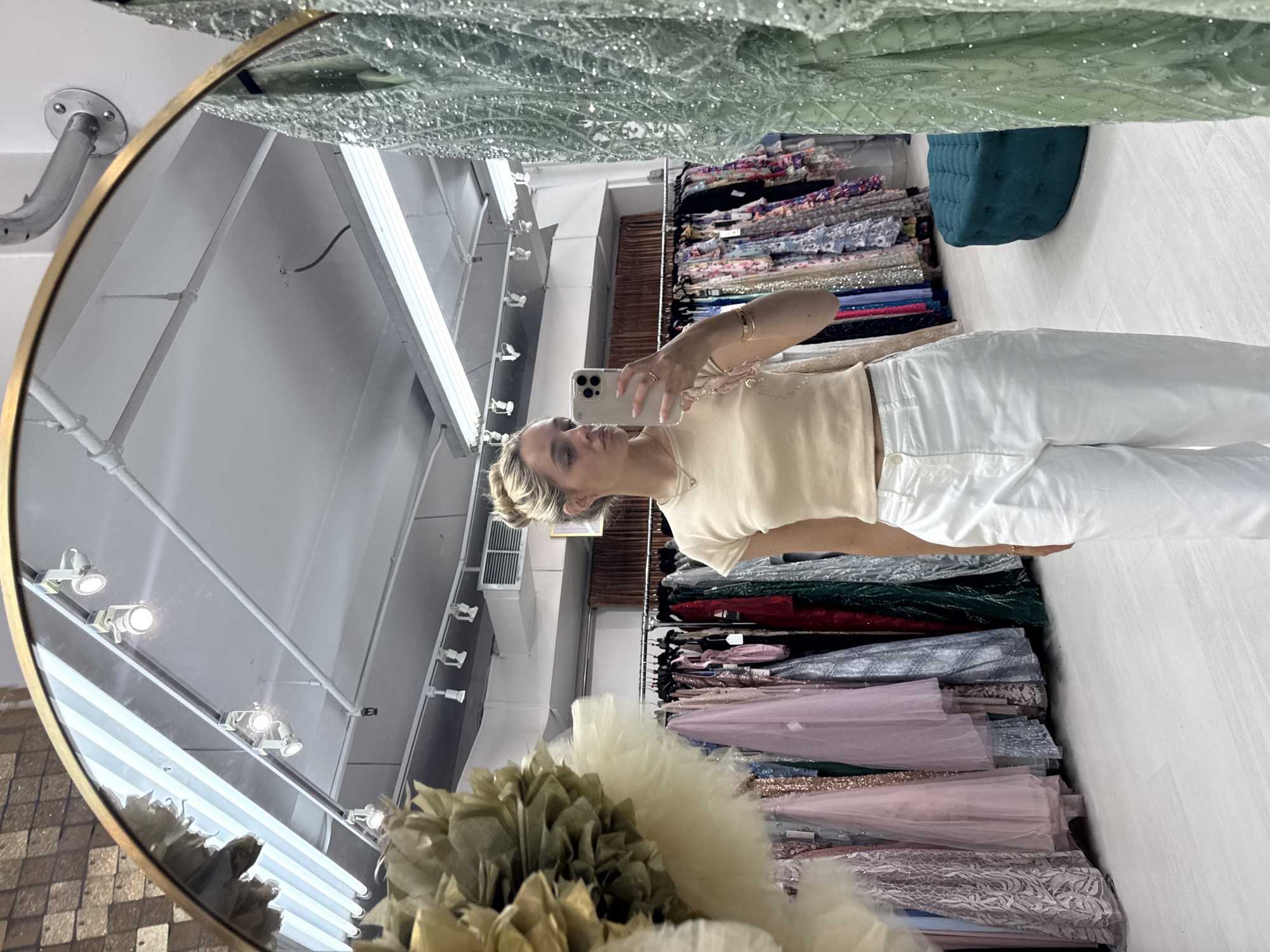 In a New York City boutique, a young Caucasian woman with blonde hair pulled into a bun, wearing a light beige crop top and white pants, is taking a mirror selfie. She is positioned in front of a rack of formal dresses, with a large decorative floral arrangement to her left and another beaded green dress to her right. The store has bright overhead lighting, a white ceiling, and a light-colored floor. A teal ottoman is visible in the background.
