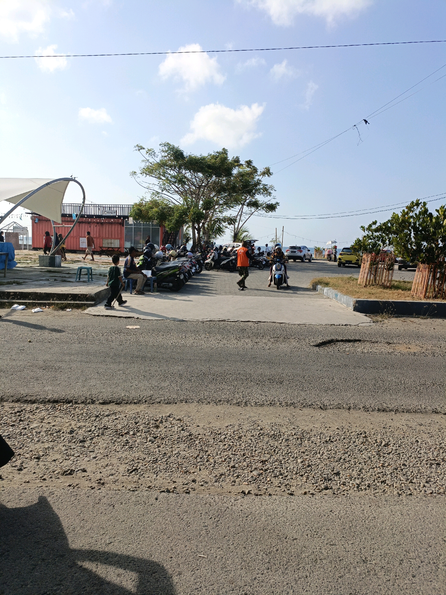 In Bulukumba, Indonesia, a street scene unfolds under a bright, partially cloudy sky. The foreground displays a cracked asphalt road, transitioning to a concrete walkway where several people are gathered and moving. To the left, a white canopy structure extends over a red shipping container building. A line of parked motorcycles is visible to the right, with a yellow car and a white SUV further down the road, suggesting a public area or possibly a parking lot near a commercial establishment or beach. A young boy in a green shirt walks across the concrete area, while other individuals are seen standing, sitting, or riding motorcycles. The environment appears to be a coastal or recreational area, with hints of water in the distance.