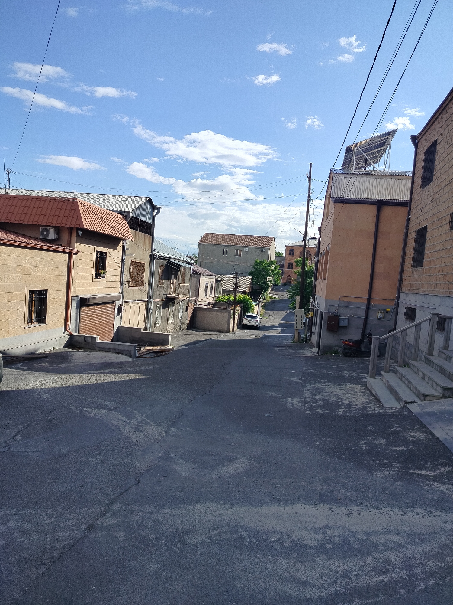 The image depicts a street in Yerevan, Armenia. The street slopes downwards, with buildings lining both sides. The buildings vary in style and construction, featuring different colors, materials, and architectural details. A car is parked further down the street. Utility lines are visible overhead, crisscrossing the sky. The sky is blue with scattered clouds. The asphalt street surface shows signs of wear and tear.