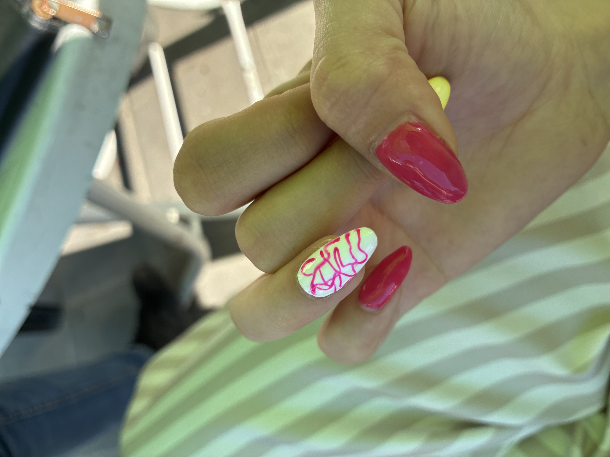 A close-up shot focuses on a woman's hand with bright pink almond-shaped nails. The index finger nail has a white base with a vibrant pink abstract design. A hint of a yellow nail is visible on the thumb. The woman is wearing a light-colored, striped fabric. The background is blurred, showing hints of furniture and what appears to be a person wearing blue jeans. The scene is indoors with soft lighting. The location is the city of Aglen, Bulgaria.