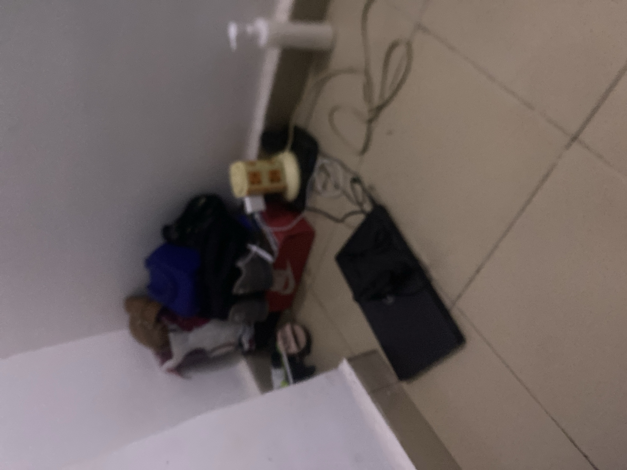 The image depicts a cluttered corner of a room with tiled flooring. In the background, against the wall, several items are piled up, including what appear to be shoes, bags, and possibly clothing. Among these items, a red shoebox with a white Nike logo is visible. To the right of this pile, there is a yellow cylindrical object, possibly a dispenser or container, resting on a black mat. Wires are scattered around this area, connecting to the yellow object. Further to the right, a white pump dispenser bottle stands upright against the wall. In the foreground, a black laptop is placed flat on the floor. The overall environment appears unorganized. The specific location in Howayi, Nigeria, cannot be determined from the visual information provided in the image.