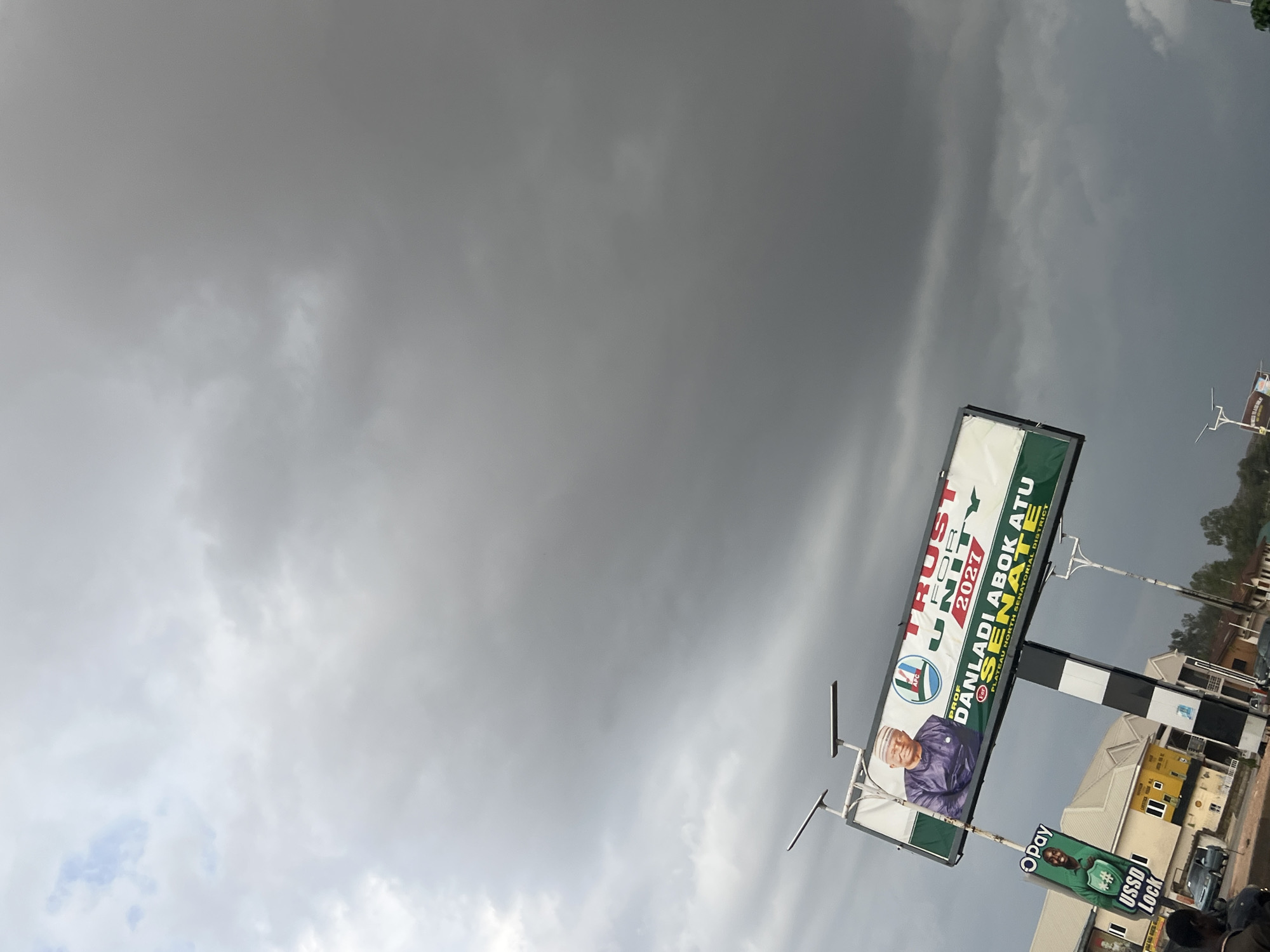 The image shows a political billboard in Jos, Nigeria. The billboard advertises Prof. Danladi Abok Atu for Senate in the Plateau North Senatorial District, under the APC (All Progressives Congress) party. Below the political ad is a smaller advertisement for Opay, a financial technology company. The sky is overcast with dark storm clouds. Buildings and vegetation are visible in the background, suggesting an urban or suburban location within Jos. There are no discernible people in the foreground of the image.