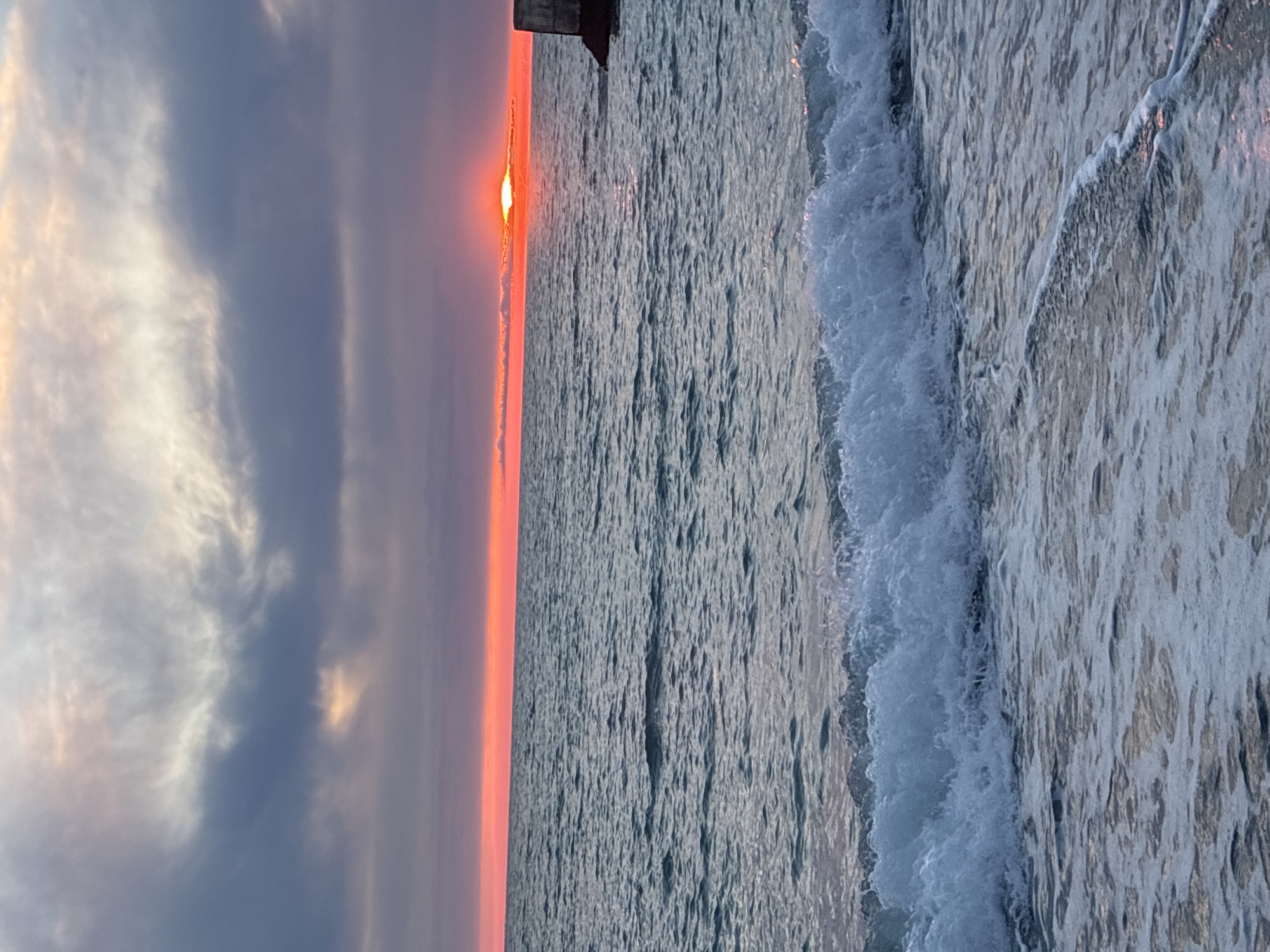 A serene sunset over the Black Sea unfolds near the Sochi coastline. The sky is a canvas of muted purples, oranges, and grays, with a band of bright orange just above the horizon where the sun dips below, casting a warm glow. Wispy clouds are scattered across the upper portion of the sky, with darker, heavier clouds closer to the horizon. The sea is a deep blue-green, with gentle waves rolling towards the shore, creating a foamy white crest on the nearest wave. A concrete structure, possibly part of a pier or breakwater, is visible on the far right of the image. The overall scene depicts a tranquil end to the day.