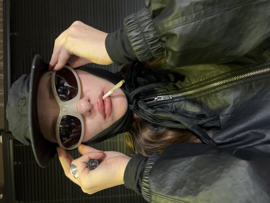A person is depicted in a close-up, chest-up vertical portrait, looking directly forward. The individual wears a dark bucket hat featuring a white "Y-3" logo on the front, positioned over a black fabric head covering that is tied around the neck. Large, light grey or off-white wrap-around sunglasses obscure the eyes, which the person holds with both hands at the temple areas. A hand-rolled cigarette with a glowing tip is held between glossy, light pink lips. Long, wavy brown hair is visible beneath the head covering and around the neck. The person is dressed in a dark, likely black, crinkled or textured jacket with a prominent metallic zipper down the front and elasticated cuffs on the sleeves. The left hand (viewer's left) wears a large, silver-toned geometric ring on the index finger and a dark ring with a red circular emblem on the middle finger. The background is a uniformly dark, featureless surface, possibly a textured wall or blinds, with subtle horizontal banding. The scene is illuminated by front-facing light, creating reflections in the sunglasses. The setting is an indoor, unidentifiable space in Tel-Aviv, Israel.
