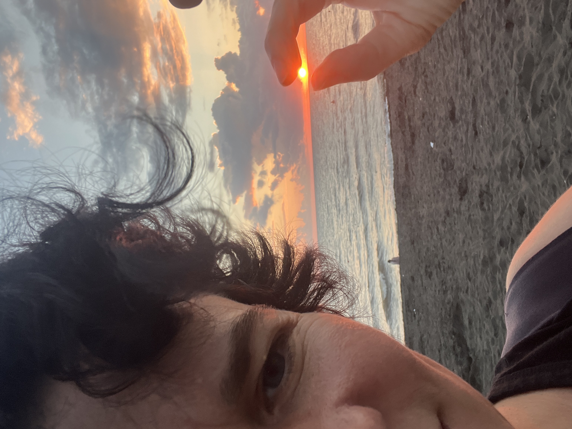 The image shows a close-up selfie of a person on a beach with dark sand. The person's face is partially visible on the left, with dark, curly hair framing their head. In the background, the sea meets the horizon under a sky filled with clouds lit by a setting sun. A hand is held up in the foreground, with fingers pinched together to seemingly frame the small, bright disc of the sun just above the waterline. The sea is calm, with gentle waves lapping the shore. In the distance, a solitary figure can be seen standing near the water's edge. The scene is characteristic of the beaches in Ureki, Georgia, known for their magnetic sand and scenic sunsets.