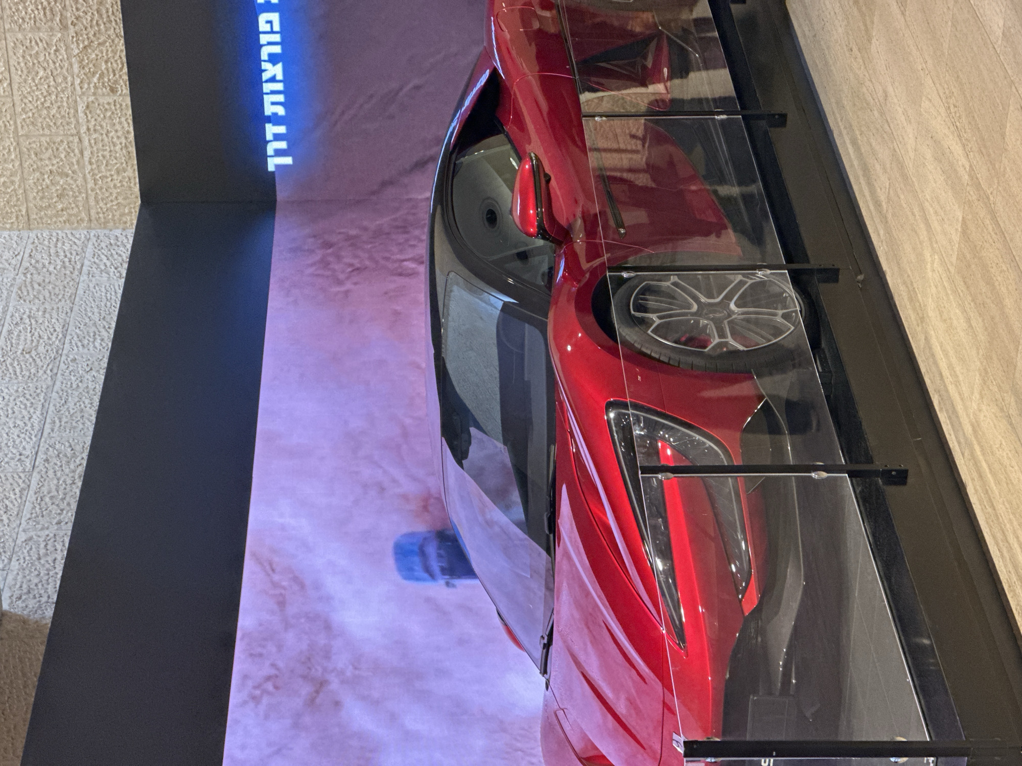 The image depicts a vibrant red sports car displayed indoors, likely in a showroom or museum. The car is positioned behind a clear barrier, possibly for protection. In the background, a large screen displays an abstract image with a sandy texture and purple fog, featuring a shadowy blue figure. Above the screen, text is visible in bright blue, appearing to be in Hebrew script, which translates to "driving miracles." The surrounding environment suggests an exhibition setting, with a textured wall above the screen and a wooden floor. The lighting is ambient, highlighting the car's glossy finish. The specific location is identified as the city of Tzafria, Israel.