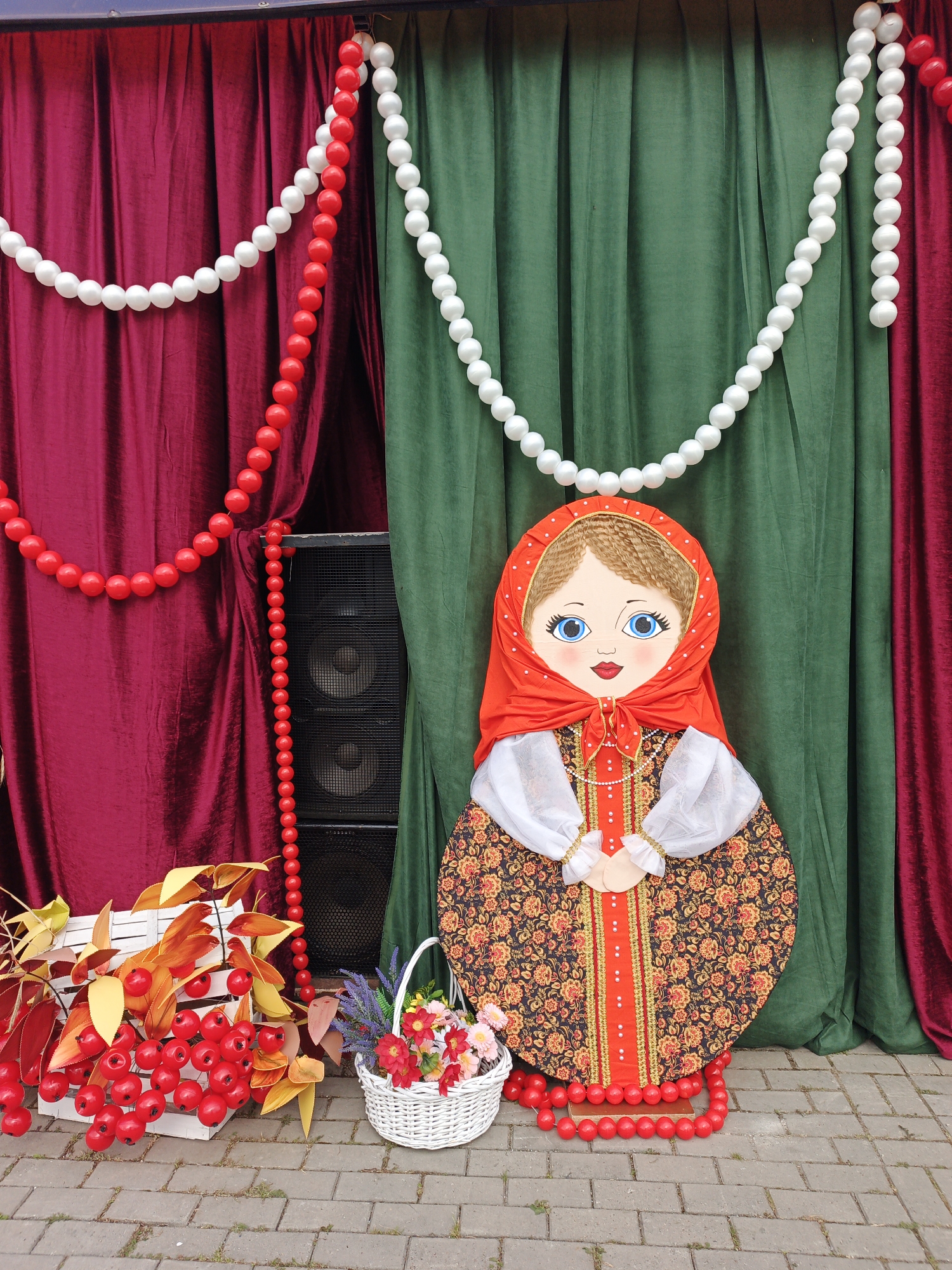 This image shows an outdoor display likely for a festival or event, featuring a large matryoshka doll as the central figure. The doll is dressed in a traditional Russian folk costume with a red headscarf, a white blouse with sheer sleeves, and a richly patterned dress in shades of red, orange, and black. The doll is adorned with red beaded necklaces.
Behind the doll, a backdrop of draped red and green velvet curtains creates a vibrant setting. White and red beaded garlands hang across the curtains, adding to the festive atmosphere. To the left of the doll, a white wooden crate is filled with red artificial berries and autumn leaves, accompanied by a white wicker basket overflowing with colorful artificial flowers. A large black speaker unit is partially visible behind the curtains, to the left of the matryoshka doll. The display is set on a cobblestone ground. The scene suggests a cultural or holiday celebration.