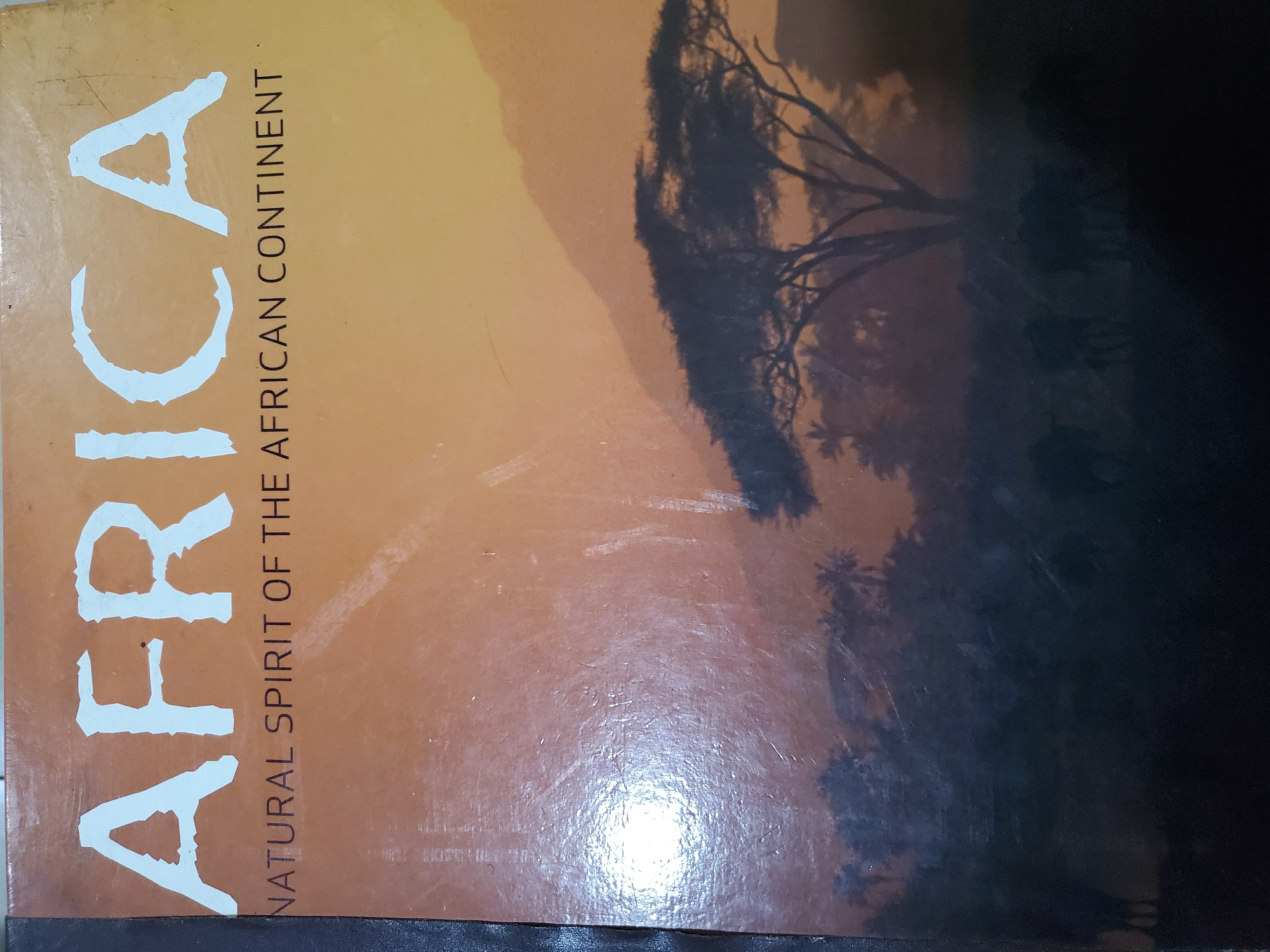 The image shows the cover of a book titled "AFRICA: NATURAL SPIRIT OF THE AFRICAN CONTINENT". The cover features a sunset over a savanna landscape with silhouetted acacia trees and mountains in the background. A herd of animals, possibly wildebeest, are visible in the foreground. The book cover is slightly worn with visible scratches. There is no visible indication of the city of Jos, Nigeria, or any specific location within it.