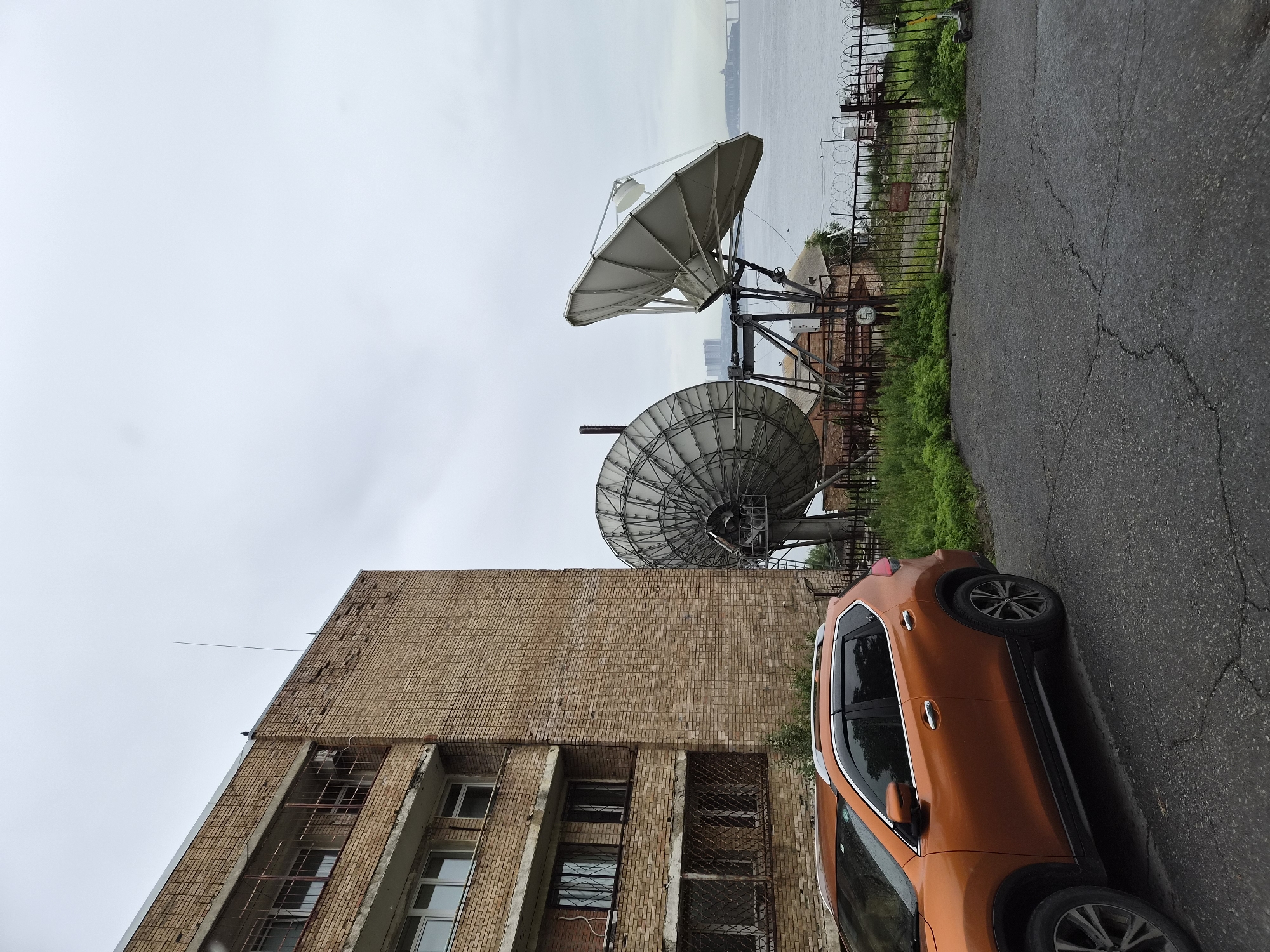 The image depicts an outdoor scene in Vladivostok, Russia, featuring a weathered, multi-story brick apartment building on the left. In front of the building, an orange SUV is parked on a cracked asphalt road. To the right of the building and behind a black metal fence with barbed wire, two large satellite dishes are mounted on a metal structure. The larger dish on the left is partially visible, while the dish on the right is fully displayed and angled upwards. Beyond the satellite dishes, a body of water, likely a bay or gulf, is visible, with a distant bridge and city skyline across it. The sky is overcast and grey. The area appears to be somewhat neglected, with overgrown grass around the fence and the building showing signs of age and disrepair. There are no people visible in the image.
