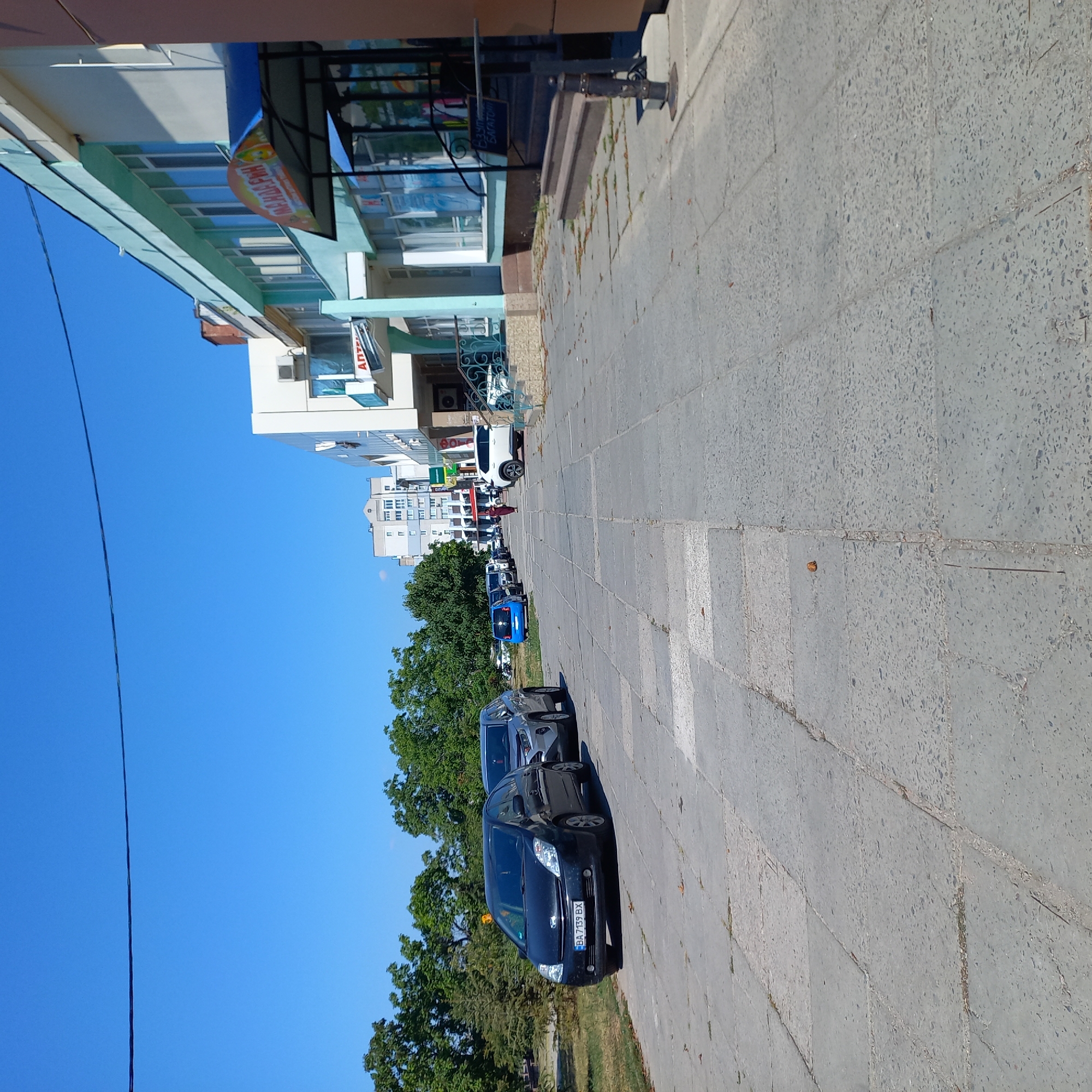 The image captures a street scene in Kropyvnytskyi, Ukraine, under a clear blue sky. A wide sidewalk paved with large square concrete slabs is in the foreground, with scattered leaves and some small patches of grass growing between the slabs.
To the left of the sidewalk, a green area with lush trees is visible, suggesting a park or a tree-lined boulevard. Several cars are parked along the street. In the immediate left foreground, a black Toyota Prius with license plate BA 7139 BX is parked, and next to it is a silver Toyota RAV4. Further down the street, other vehicles are parked, including a small blue car and several others further in the distance.
On the right side of the street, a light-colored, multi-story building lines the sidewalk. This building appears to house commercial establishments, with awnings and signage visible. One awning displays the word "МАНДАРИН" in colorful letters. Another sign is partially visible with the Cyrillic word "АПТЕКА," indicating a pharmacy. The architecture suggests a relatively modern urban setting. A person in a dark red dress is walking away from the viewer down the sidewalk towards the distant buildings. The overall impression is a typical sunny day in a city with a mix of residential and commercial buildings and parked vehicles.