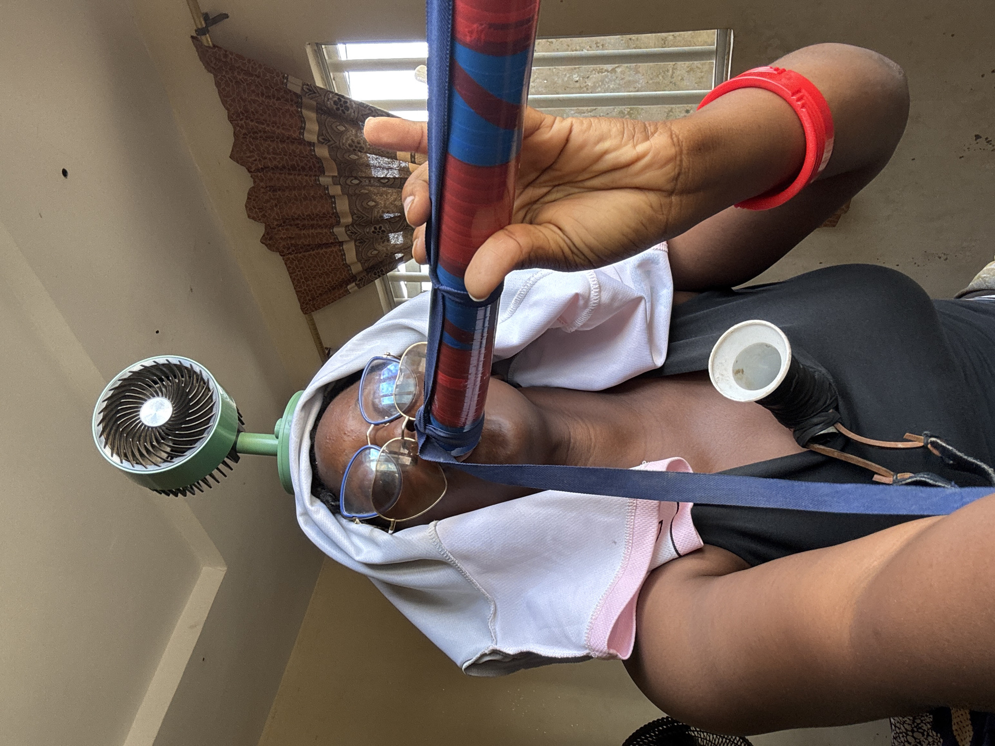 A medium shot captures a woman in Jawu, Nigeria, holding a red and blue striped tube to her mouth. She is wearing glasses and a white towel draped over her head, with a small fan attached to a stand positioned above her. The woman has a black tank top on and a red bracelet on her right arm. A white cup is attached to a strap around her neck. The background includes a window with a brown patterned curtain. The overall impression is of an attempt to find relief from heat.