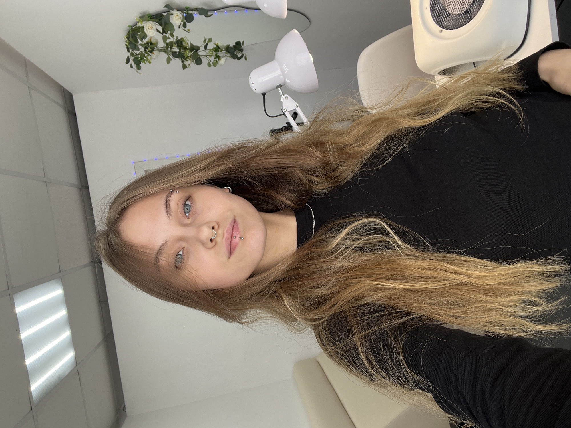 A young adult female is depicted in a close-up selfie taken indoors. She has long, light brown and blonde hair, blue eyes, and multiple facial piercings, including a silver septum ring, a silver labret stud below her lower lip, a small silver barbell piercing above her left eyebrow, and a small silver hoop earring in her left earlobe. She wears a black long-sleeved top and a thin silver chain necklace. The background features white walls and a white ceiling with recessed fluorescent panel lighting. In the upper right corner, a decorative arrangement of artificial green foliage and white roses is visible, intertwined with blue string lights. To the subject's right, a white adjustable desk lamp and a white nail dust collector with a circular vent are present on a surface, alongside a white chair. A cream-colored armchair is partially visible on the left. The scene is brightly lit by artificial light. Location: Фоки, Russia.